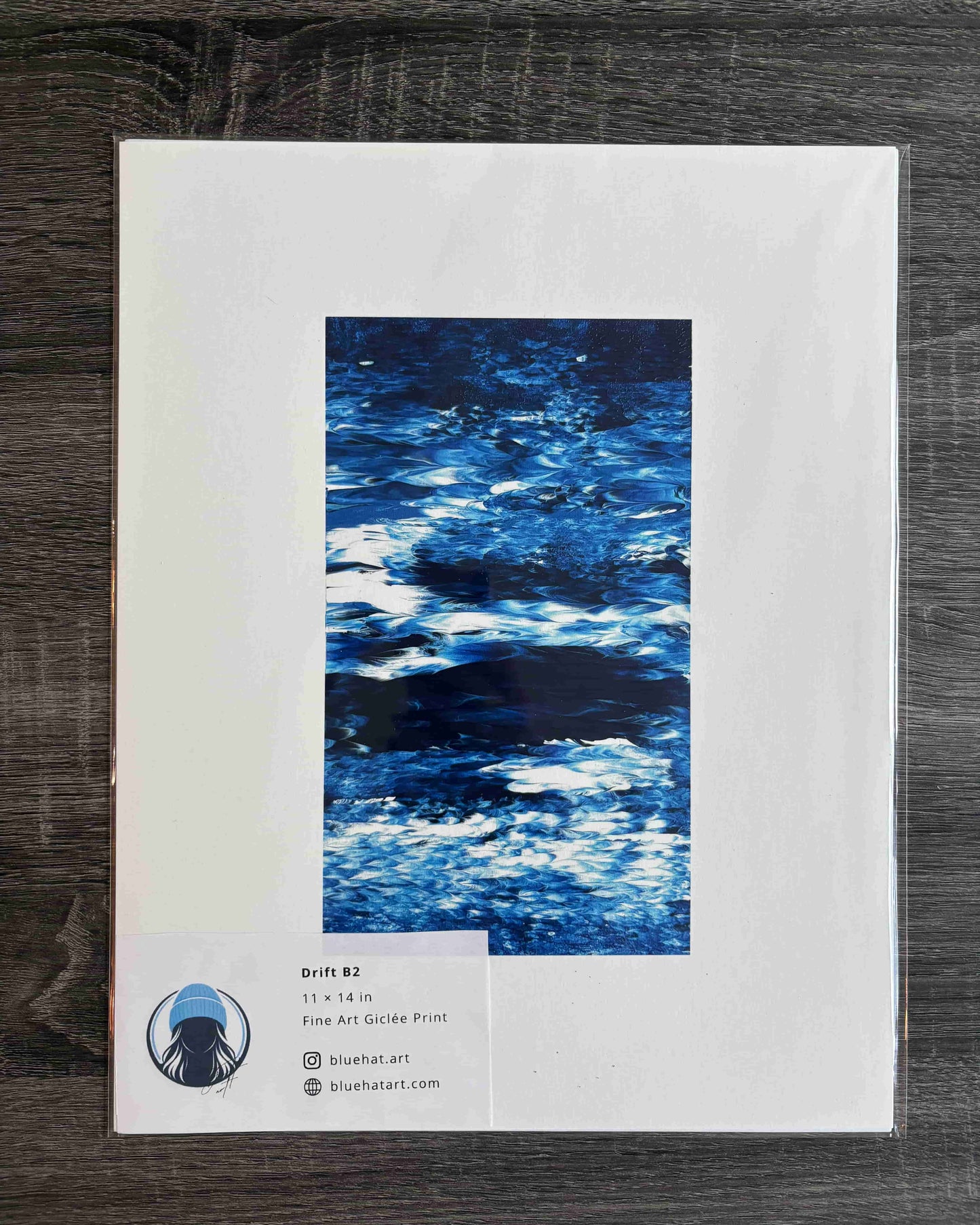 An art print package lying on a dark wood surface. The package has a large white border with a central vertical blue abstract water-like artwork and a small label at the lower left showing the brand/site