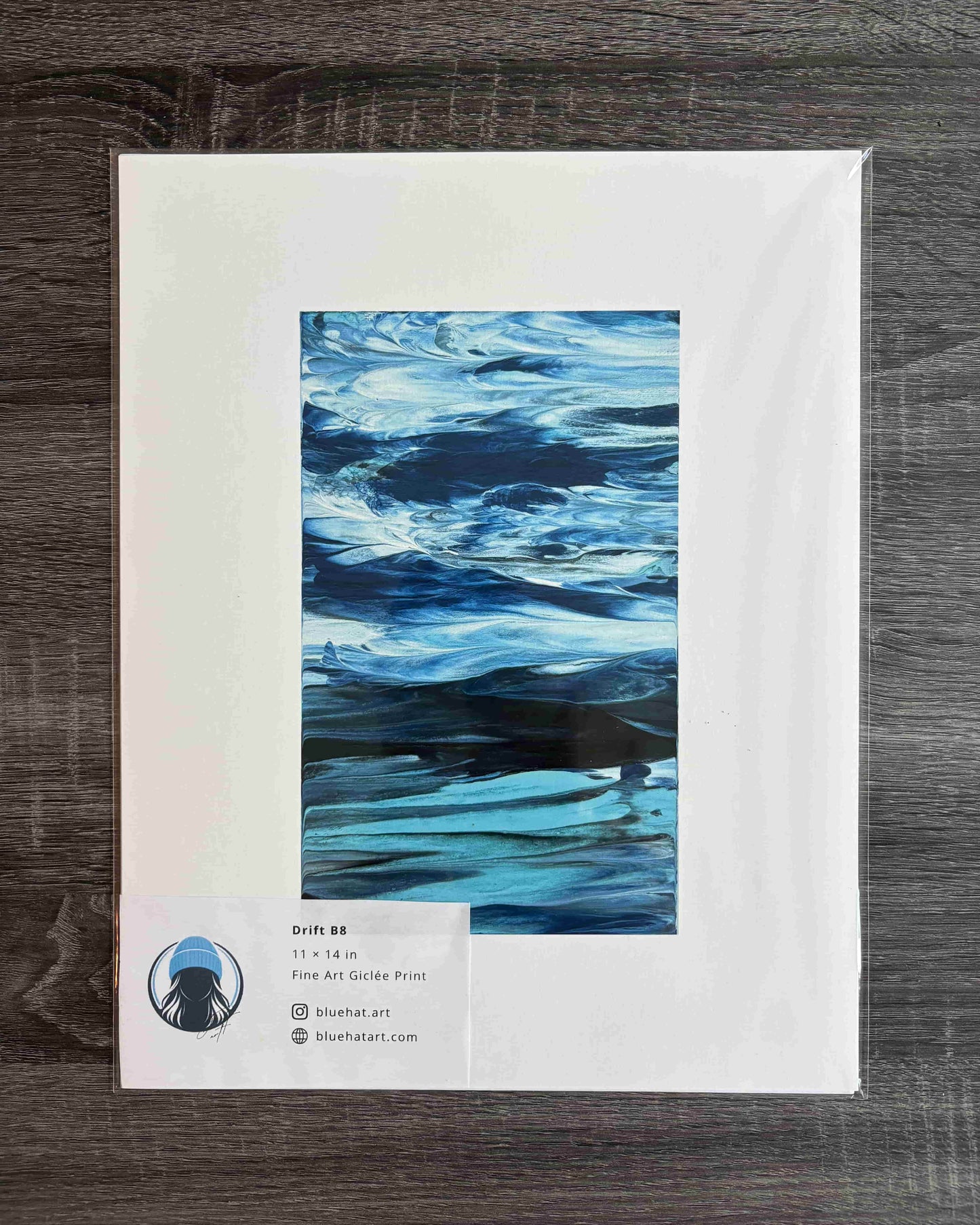A framed or matted art print showing an abstract blue water-like composition, placed on a dark wood surface. The print has a white border and a small label at the bottom left with text
