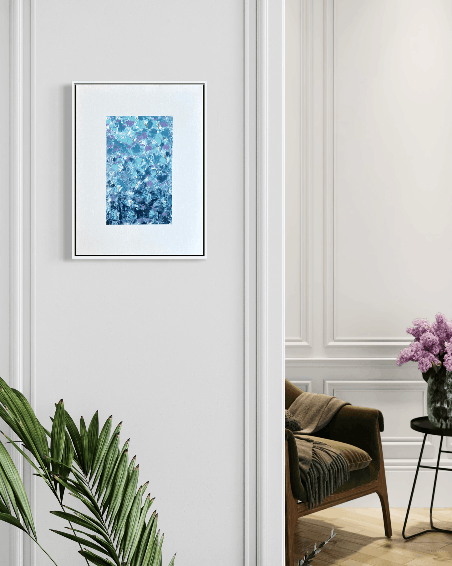 A framed abstract art print featuring a textured blue and teal floral pattern, displayed on a white paneled wall in a styled interior room with a green armchair and a plant.