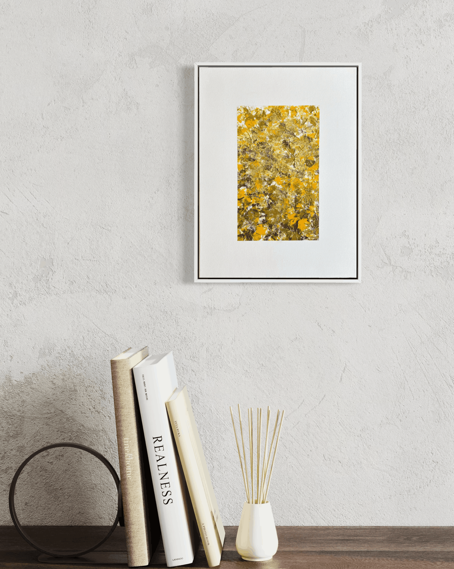 A framed abstract art print featuring a textured yellow and gold pattern, displayed on a light gray textured wall above a wooden shelf with books and a reed diffuser.
