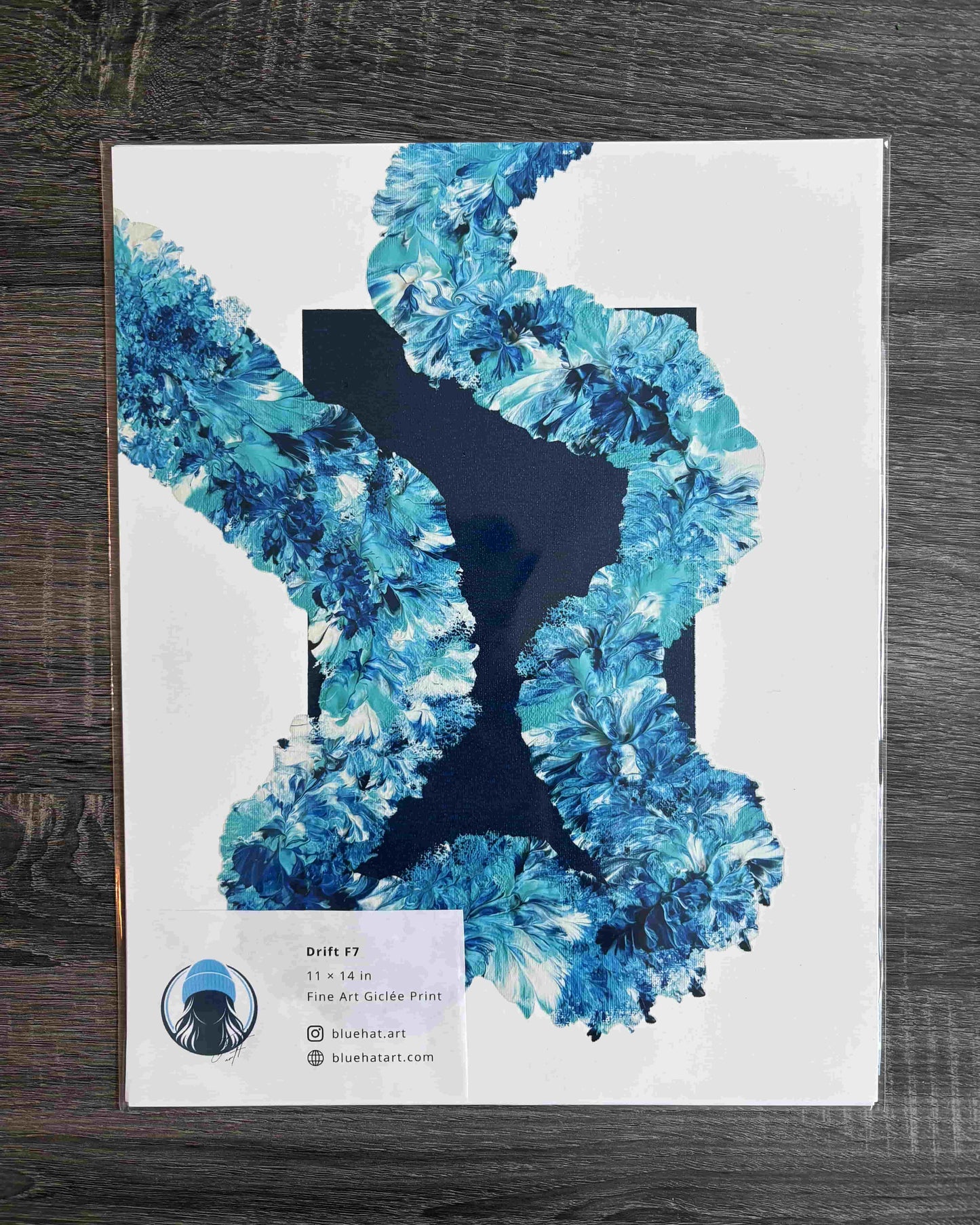 An art print titled 'Draft 2' featuring a white background with a large blue and teal floral or feather-like graphic shape, photographed on a dark wood surface. A small label at the bottom left shows the artist name 'Skeletart' and website 'skelartart.