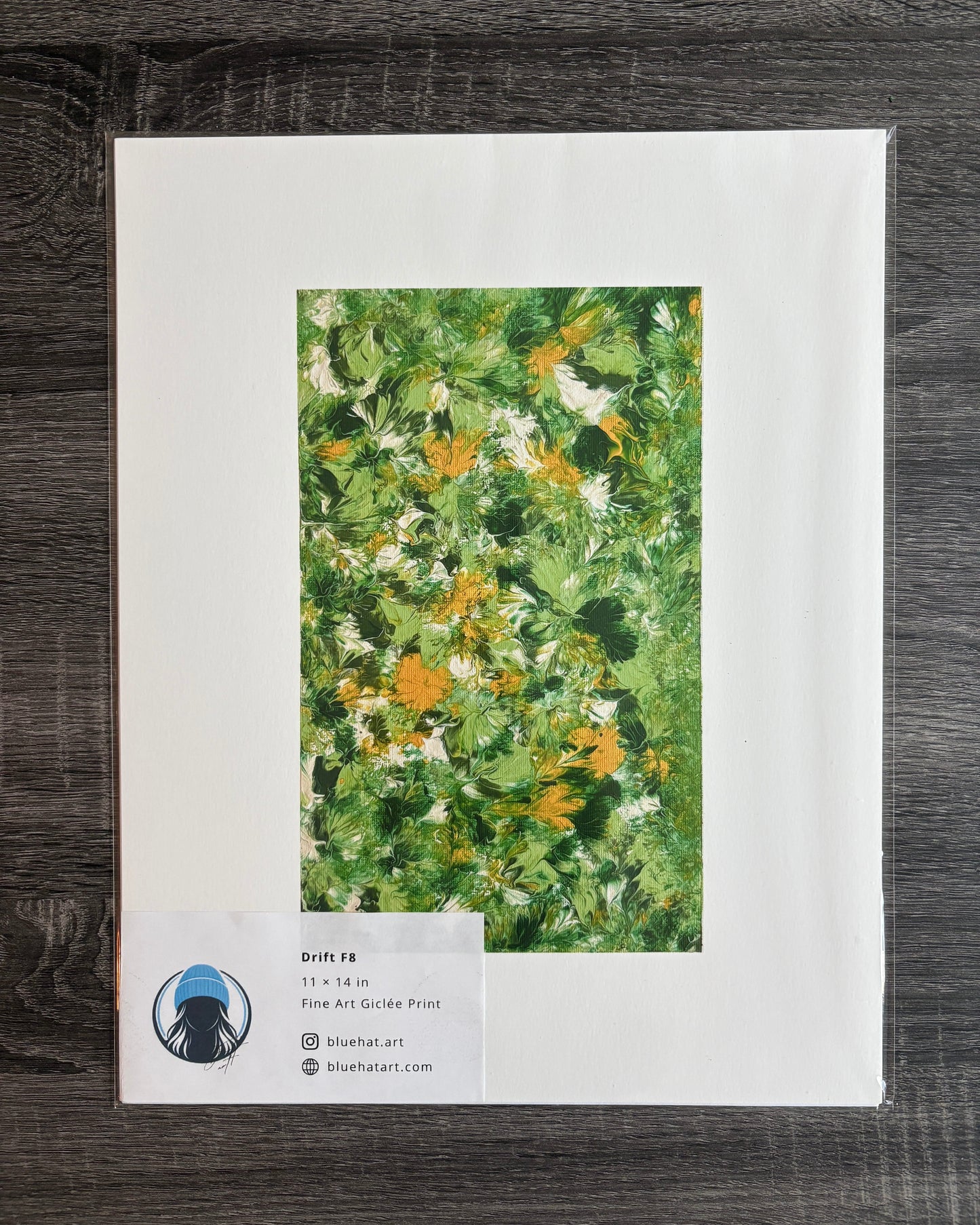 A flat-lay photograph of a matted art print on a dark wood background. The print features a textured green and yellow floral painting surrounded by a wide white mat border.