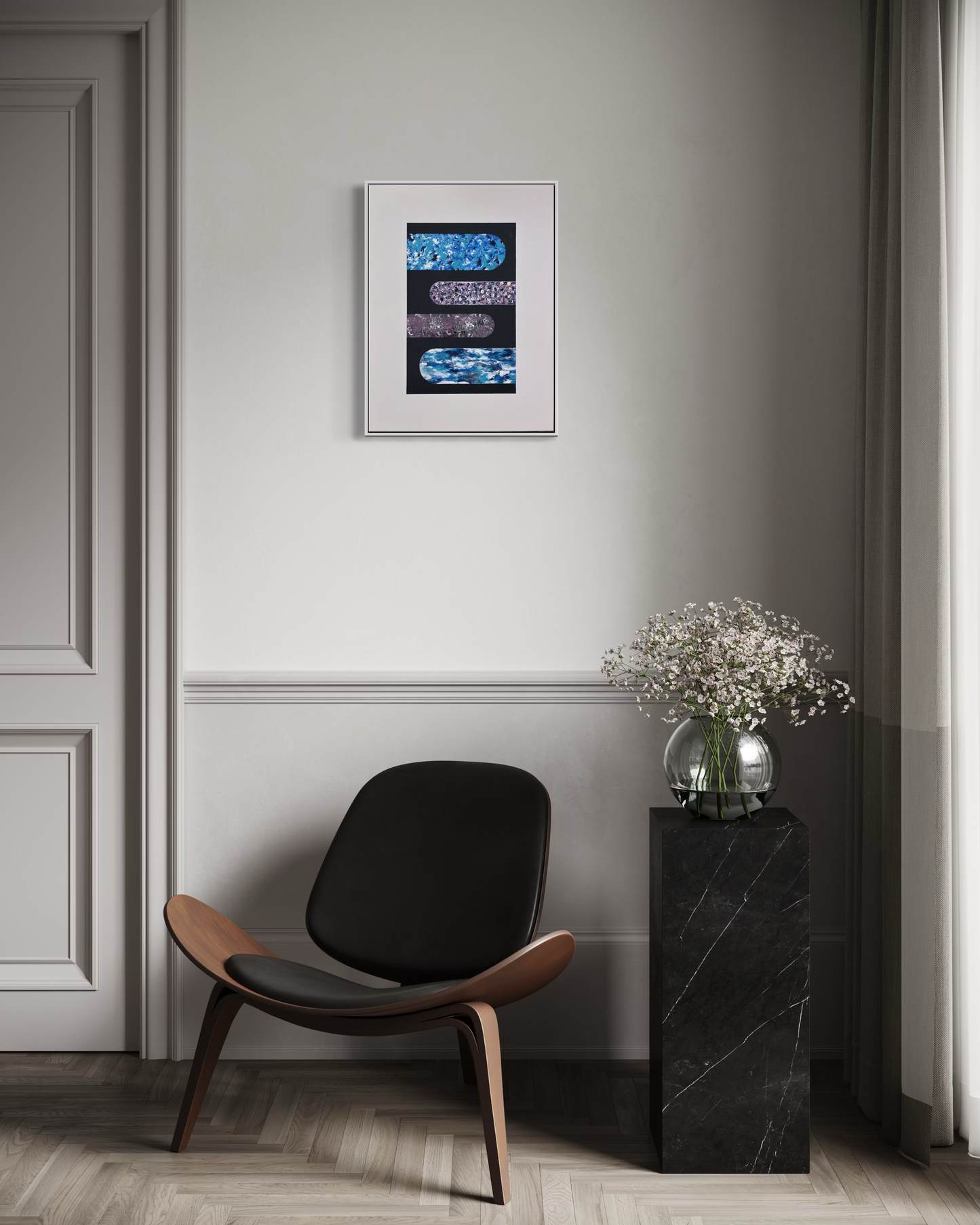 A framed abstract art print with blue, black, and purple patterns hangs on a light gray wall above a black lounge chair and a black marble side table with a glass vase of white flowers.