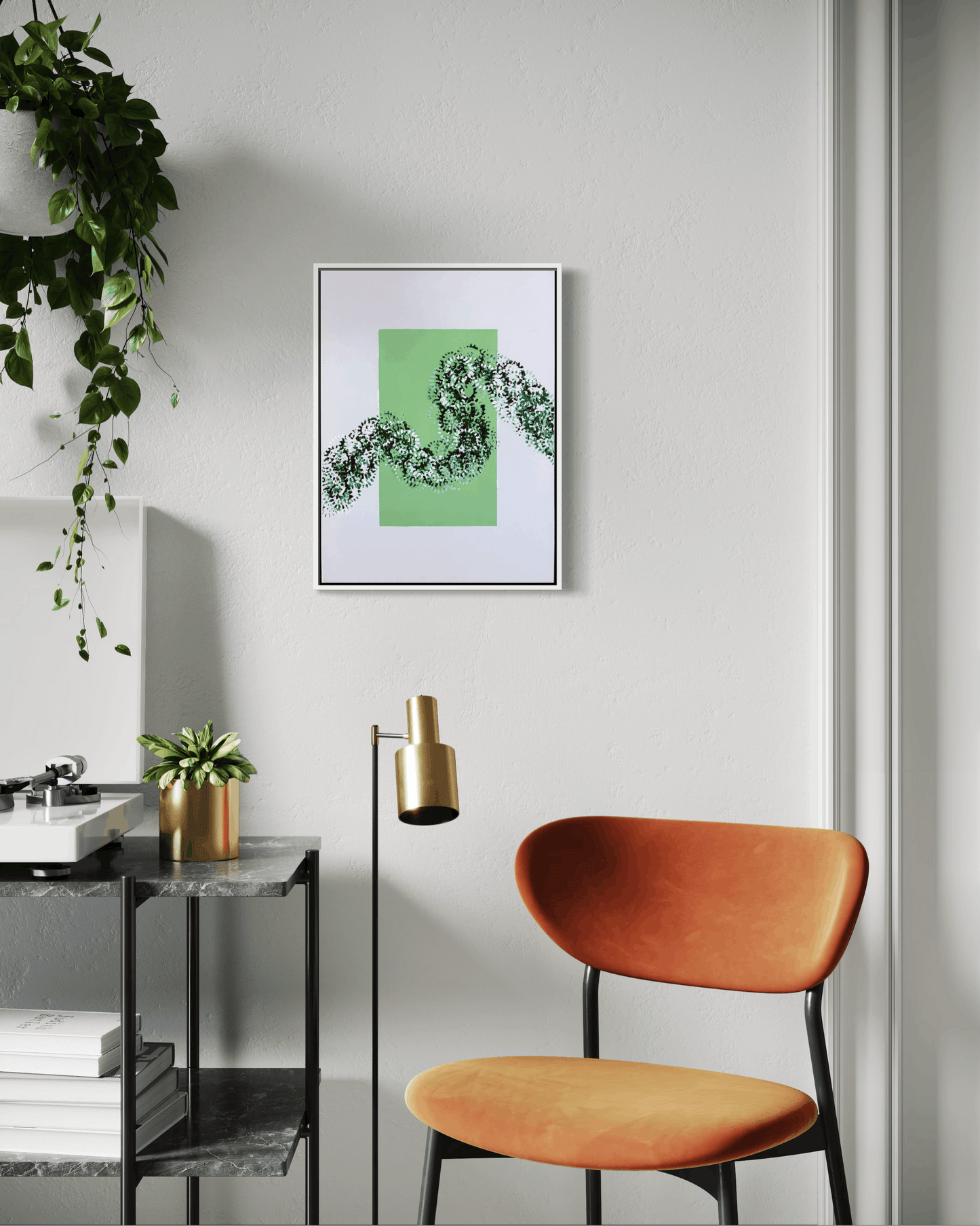 A framed abstract art print featuring a green square and a black leafy vine pattern, hanging on a light gray wall in a modern room setting with an orange chair and a side table.