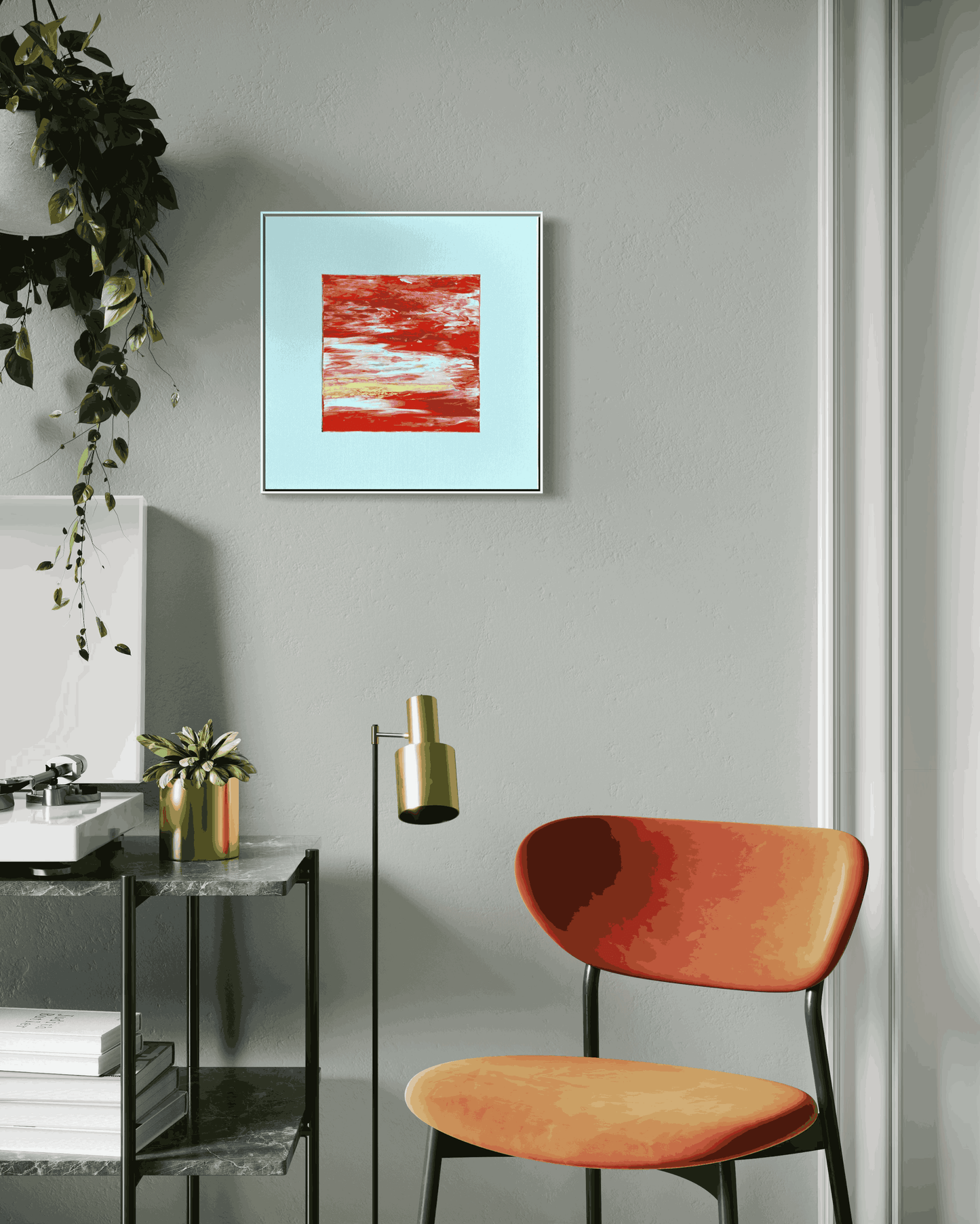 A framed abstract art print featuring textured red and white brushstrokes on a light blue background, displayed on a gray wall in a modern room setting with an orange chair and a black side table.