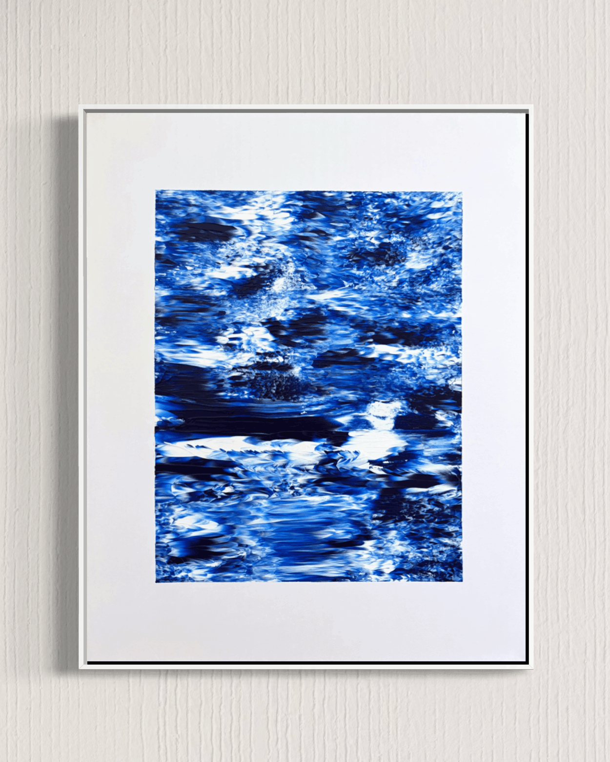 A framed abstract art print featuring a textured blue and white pattern resembling water ripples, presented in a white frame against a light gray textured wall.