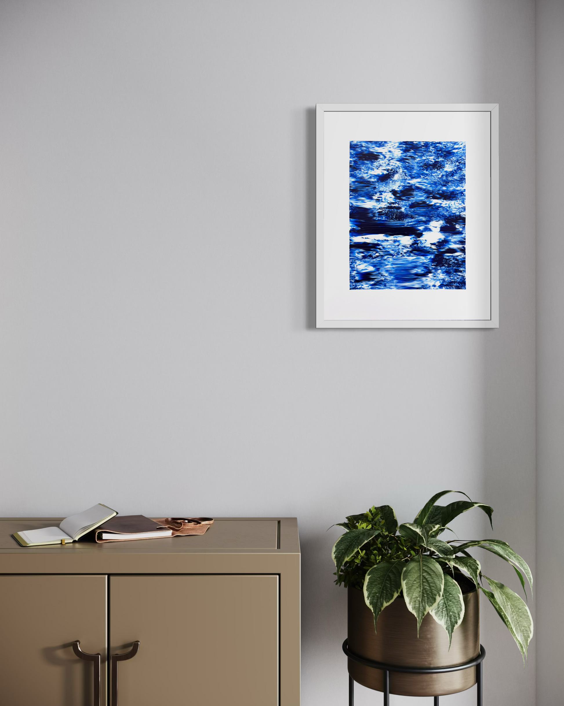 A framed abstract art print featuring vibrant blue and white brushstrokes, displayed on a light gray wall above a beige cabinet with a potted plant nearby.