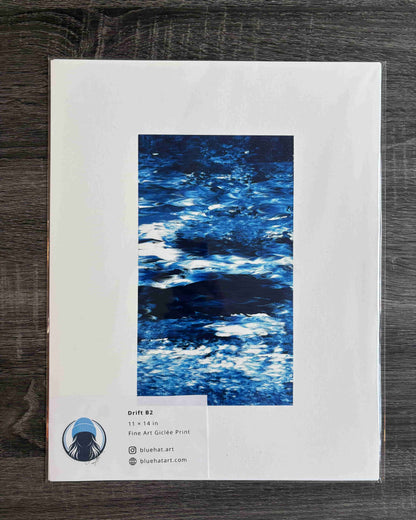 An art print package lying on a dark wood surface. The package has a large white border with a central vertical blue abstract water-like artwork and a small label at the lower left showing the brand/site