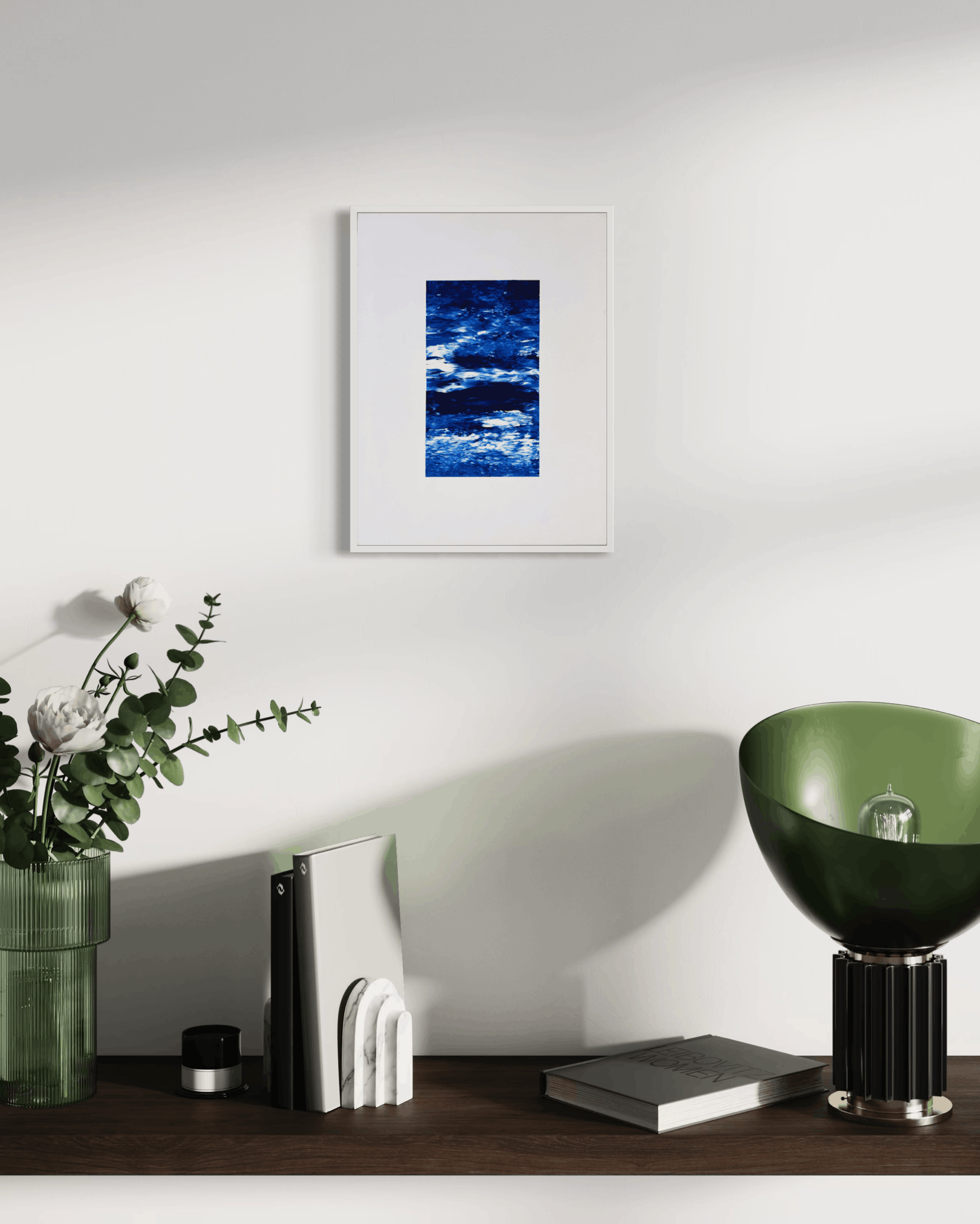 A framed abstract blue and white art print hangs on a white wall above a dark wooden shelf styled with a green vase of flowers, white bookends, a green bowl lamp, and books.