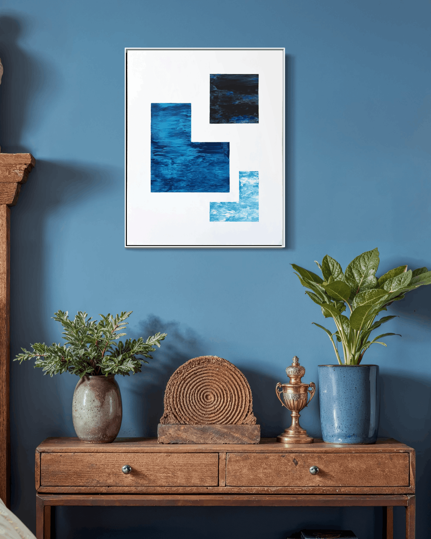 A framed abstract art print with blue and black geometric shapes on a white background, hanging on a blue wall above a wooden console table with plants and decorative objects.