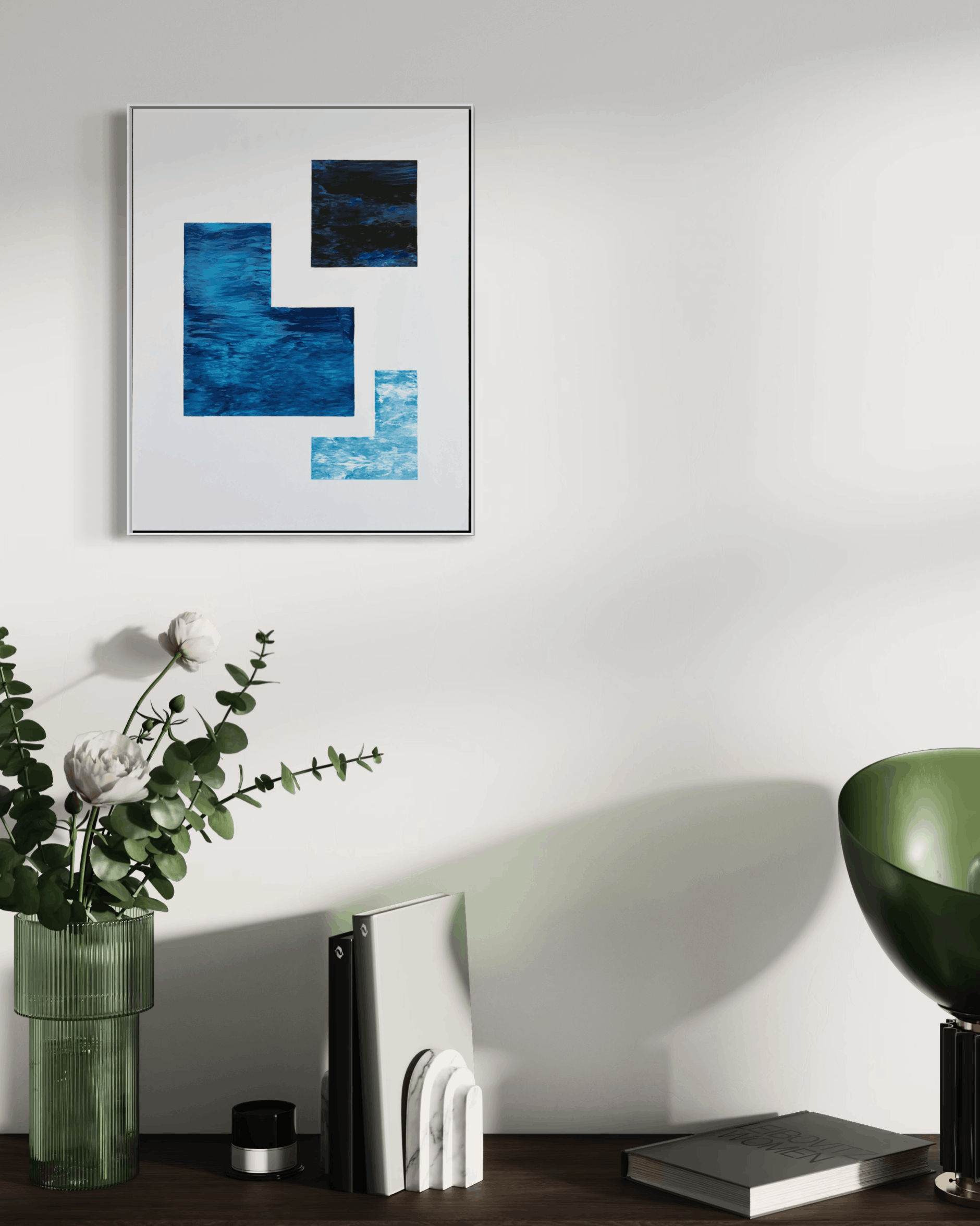 A framed abstract art print with blue and black geometric shapes hanging on a white wall above a styled desk with a vase of flowers, books, and a green lamp.