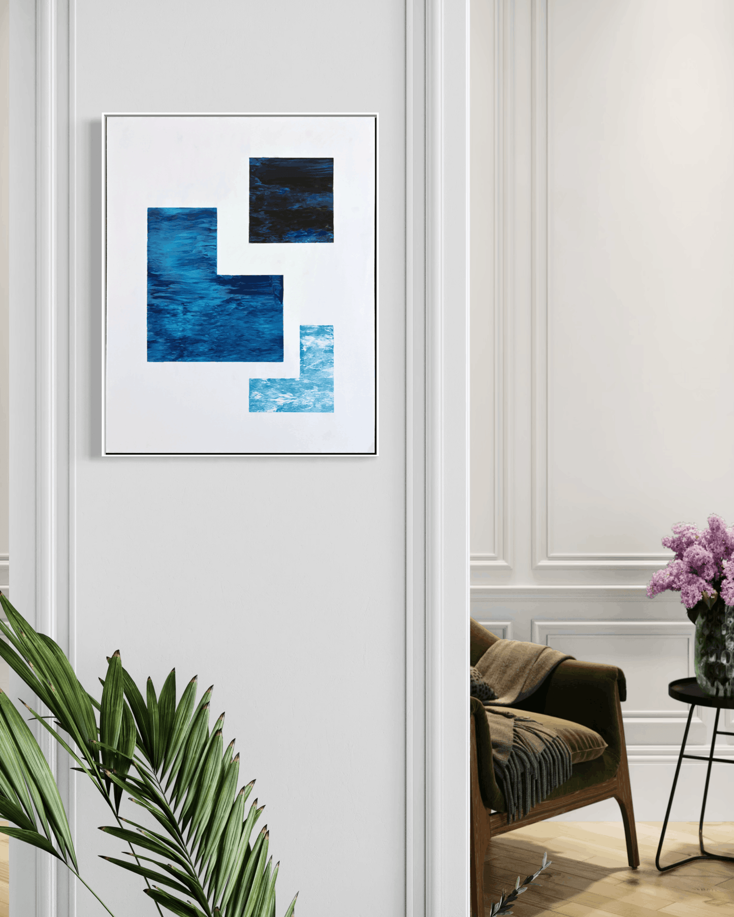 A framed abstract wall art print featuring geometric blue and navy blocks on a white background, displayed on a white paneled wall in a styled interior with a chair, side table, flowers, and a plant visible.