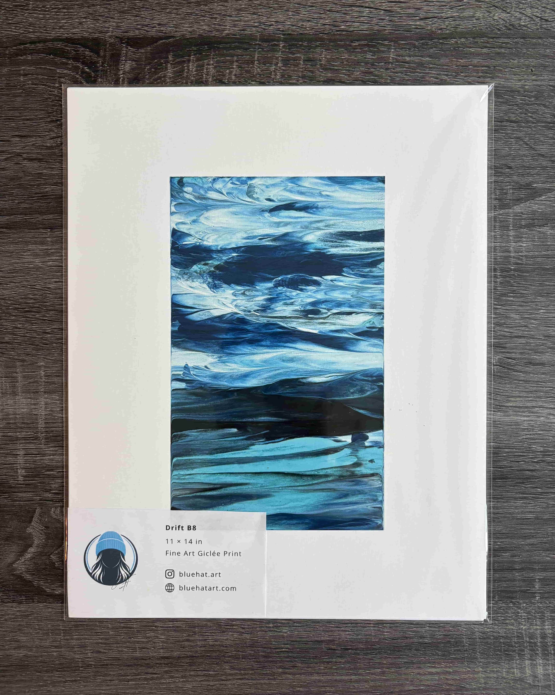 A framed or matted art print showing an abstract blue water-like composition, placed on a dark wood surface. The print has a white border and a small label at the bottom left with text 