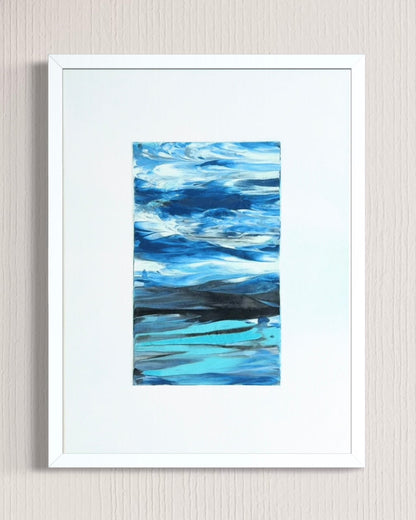 A framed abstract landscape painting featuring shades of blue, teal, and white, presented in a white frame with a wide white mat against a light beige textured wall.