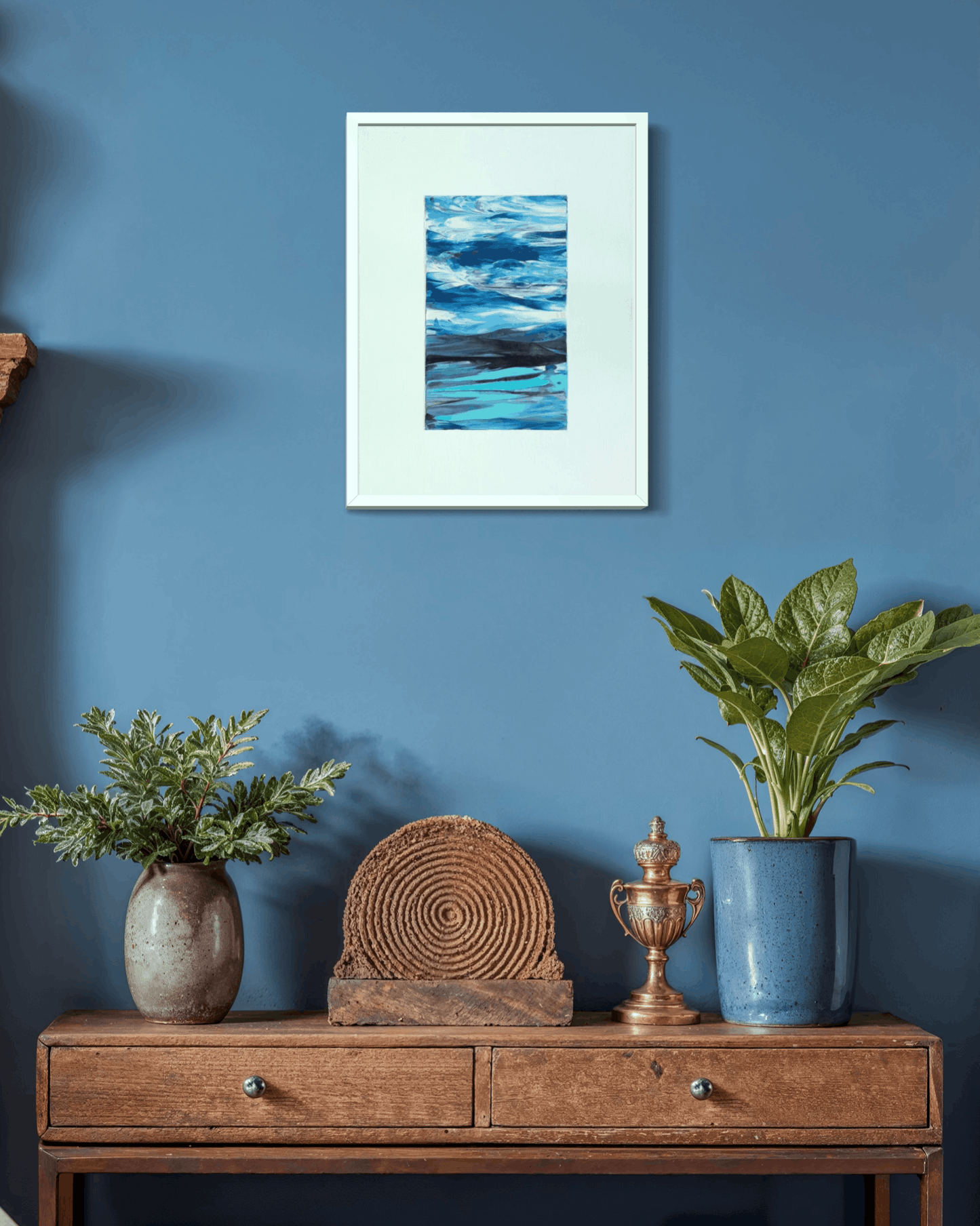 A framed abstract landscape art print featuring blue and white water waves, displayed on a blue wall above a wooden console table with plants and decorative objects.