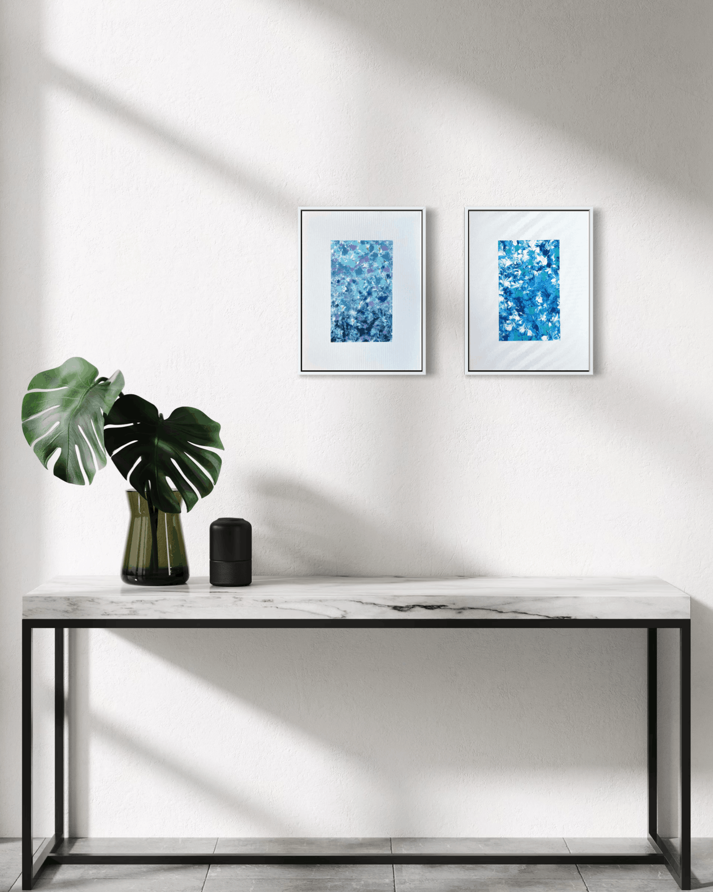 Two framed blue abstract art prints hang on a white wall above a marble console table with a black metal frame. A green monstera plant in a dark glass vase and a small black cylindrical object sit on the table, with soft diagonal sunlight casting shadows across the wall and tabletop.