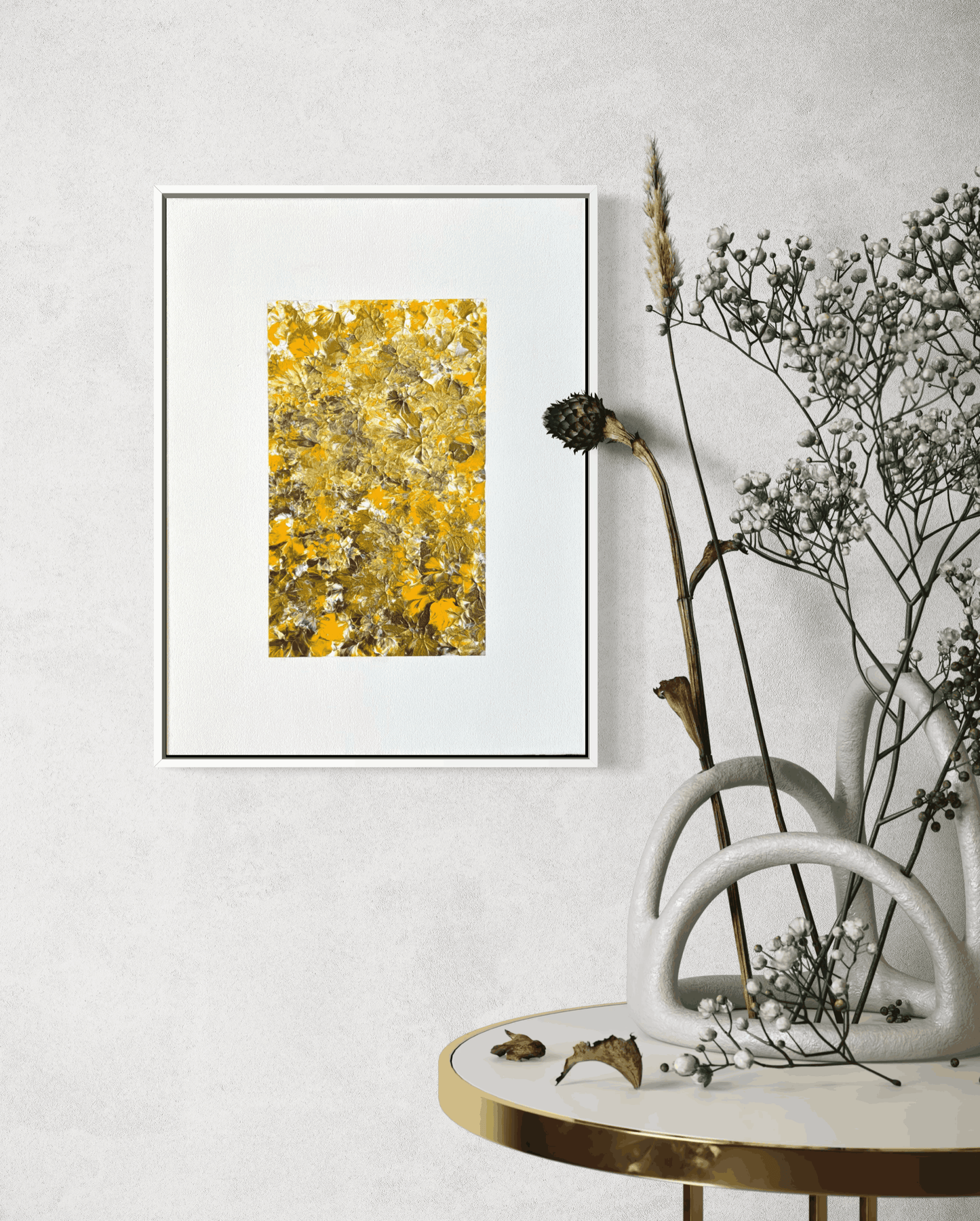 A framed abstract art print featuring a textured yellow and gold pattern on a white mat, displayed on a textured light gray wall above a round table with dried botanicals in a white ceramic vase.