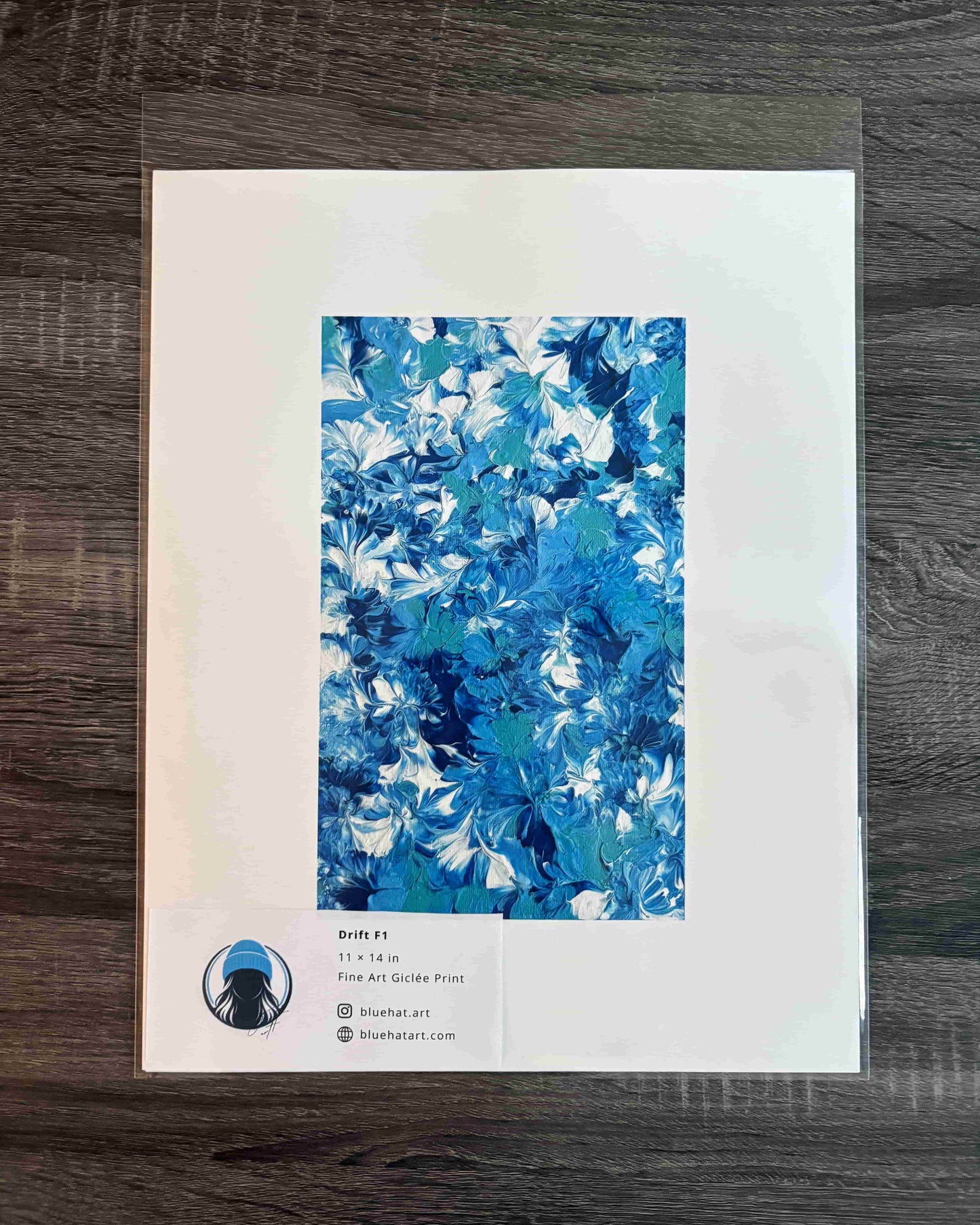 An art print featuring an abstract blue and white marbled pattern, presented on a white paper sheet with a clear protective sleeve, set against a dark wood background.