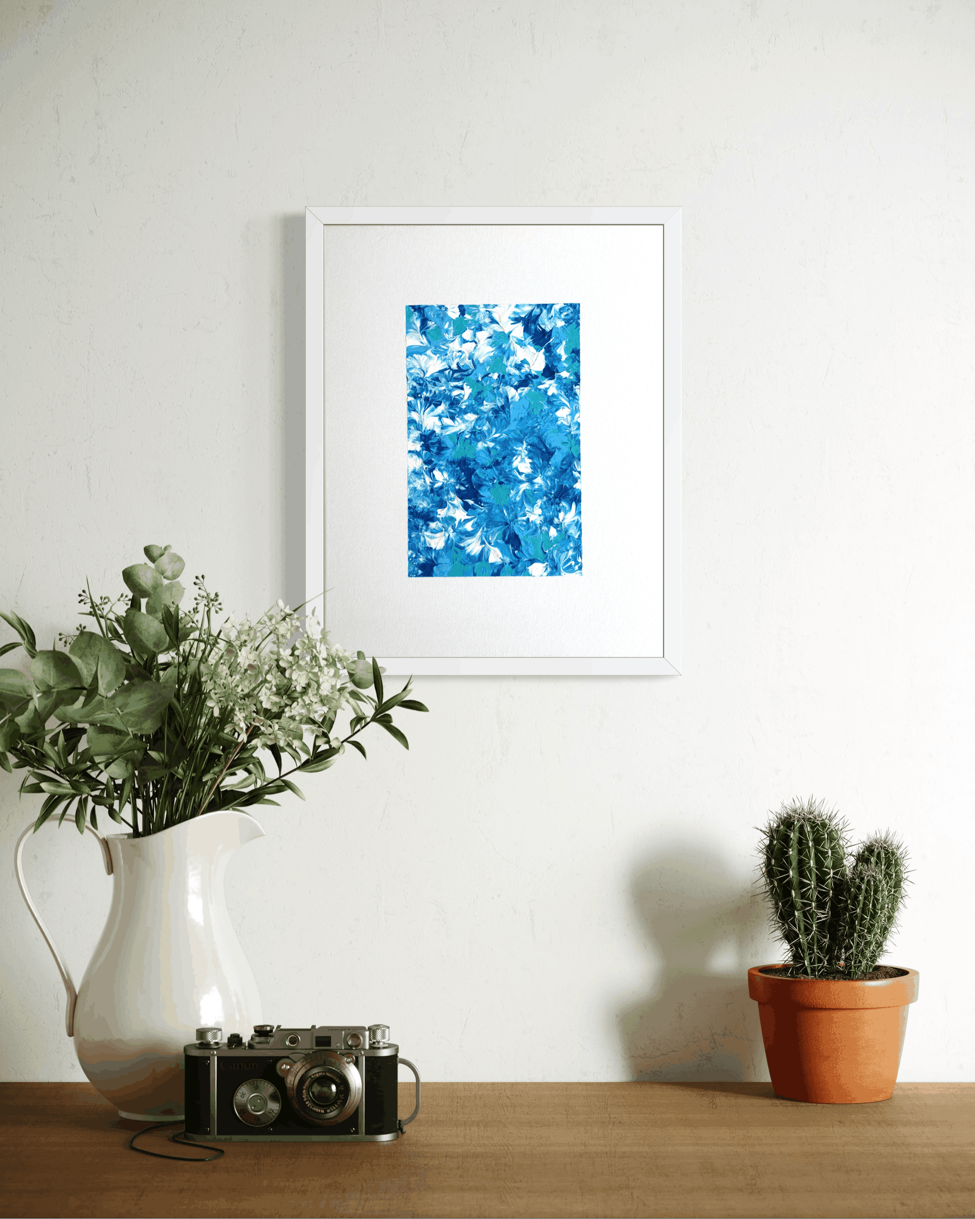 A framed abstract art print featuring vibrant blue and white brushstrokes, displayed on a white wall above a wooden table styled with a white pitcher of greenery, a vintage camera, and a small potted cactus.