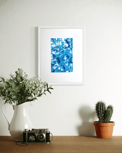 A framed abstract art print featuring vibrant blue and white brushstrokes, displayed on a white wall above a wooden table styled with a white pitcher of greenery, a vintage camera, and a small potted cactus.