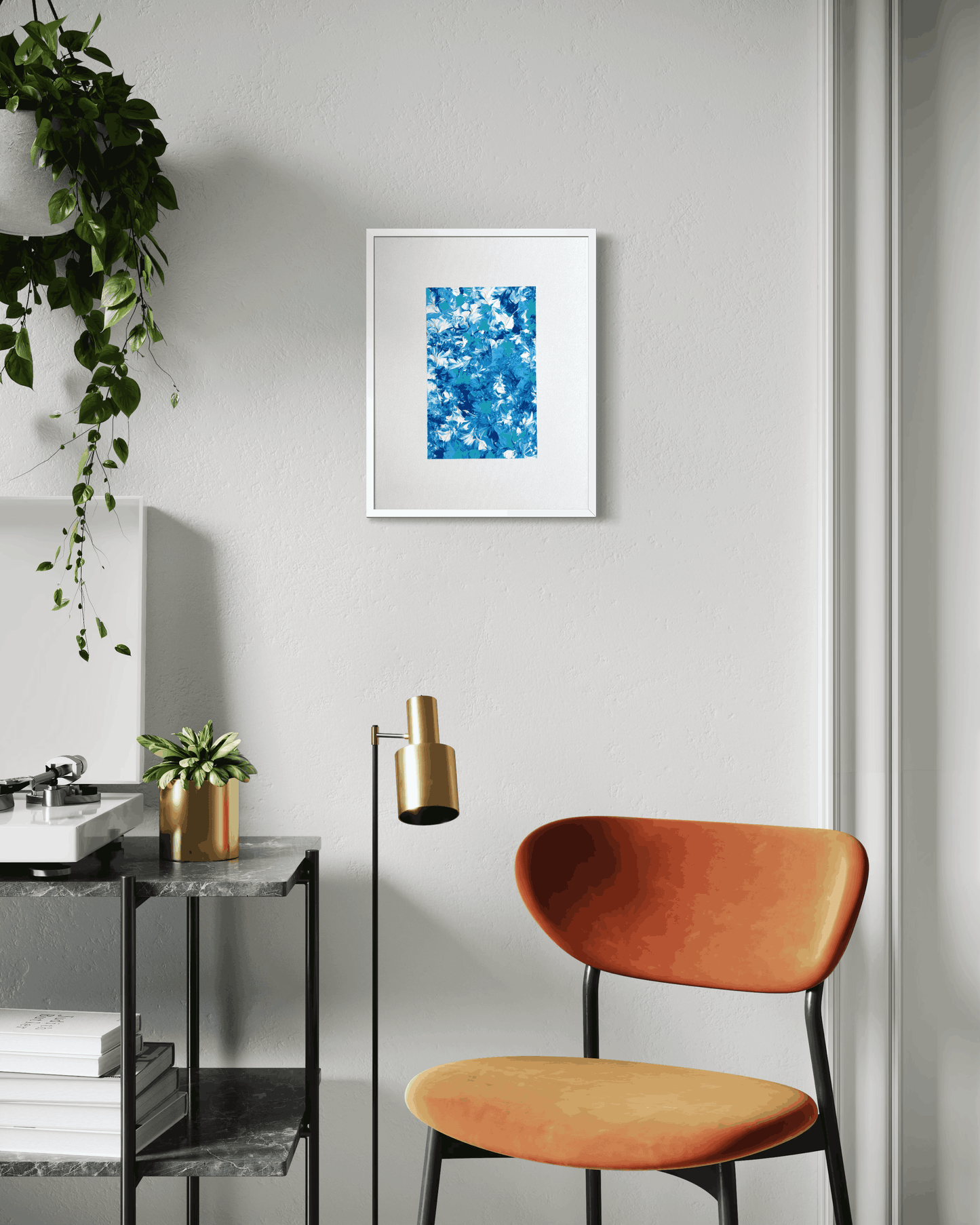 A framed abstract art print featuring a vibrant blue and white pattern, displayed on a light gray wall in a modern room setting with an orange chair and a side table.