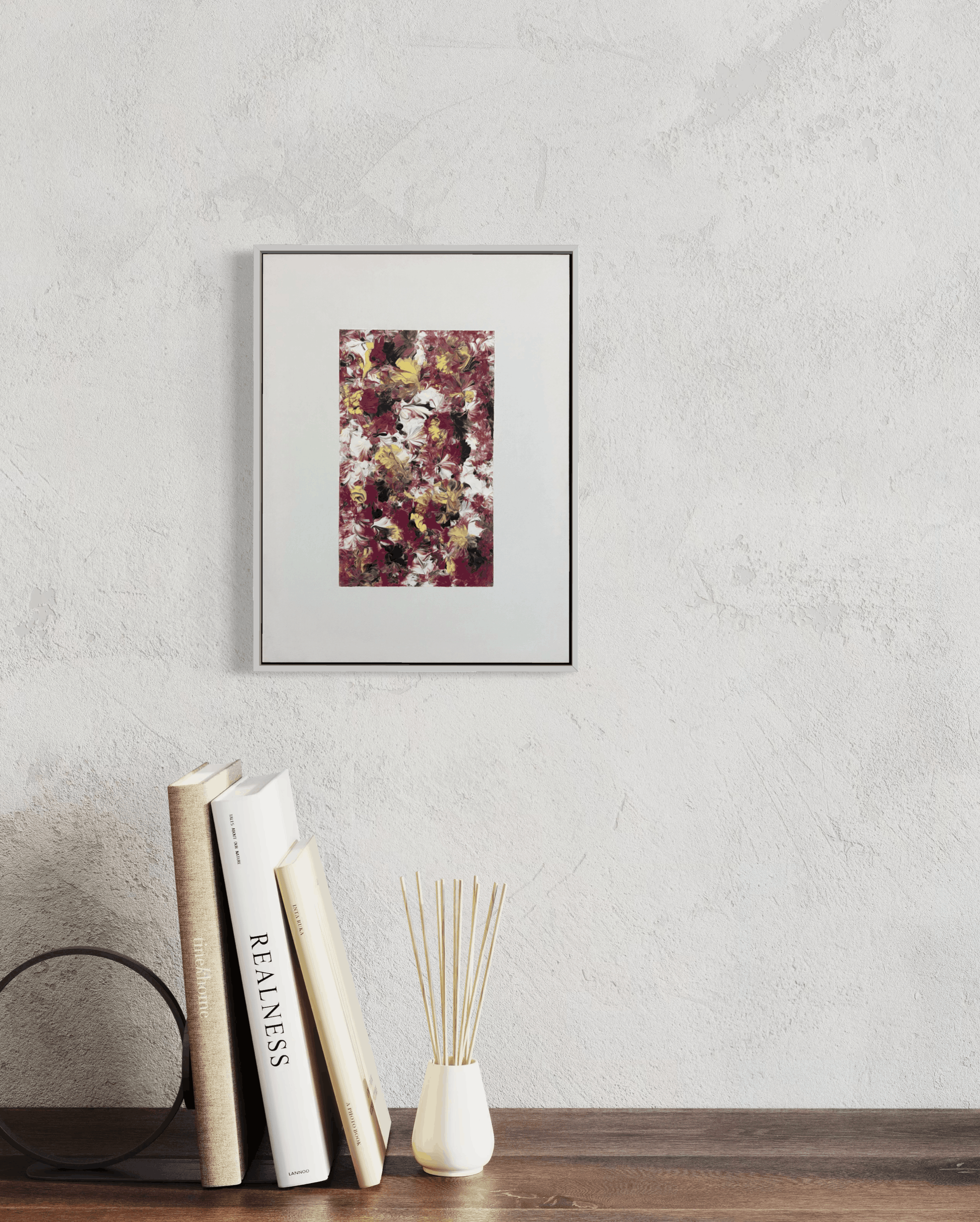 A framed abstract art print with a white mat hangs on a textured light gray wall above a wooden table styled with books, a black circular object, and a small white vase with reeds.