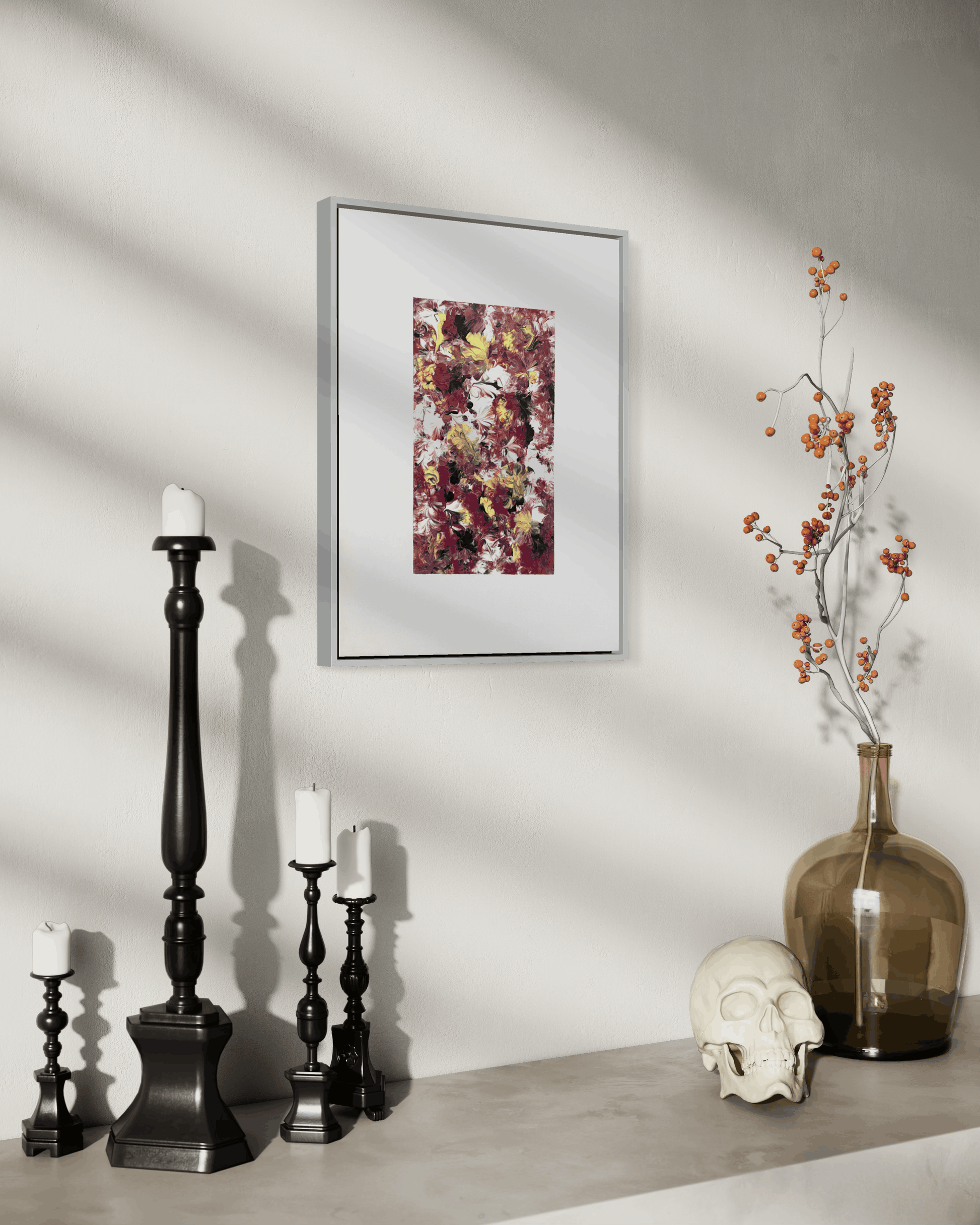A framed abstract art print with a white mat and thin silver frame hangs on a light gray wall above a console table styled with black candlesticks, a brown glass vase with dried branches, and a white skull.