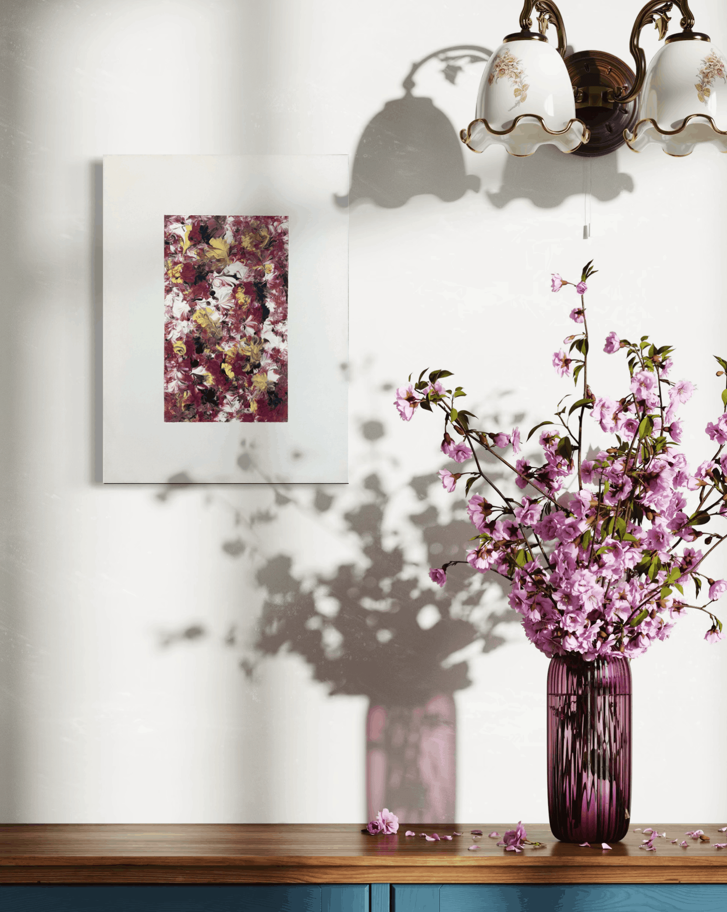 An abstract floral art print with purple, pink, and gold tones is displayed on a white wall above a wooden console table. A purple glass vase filled with pink blossoms sits on the table, casting a shadow on the wall.