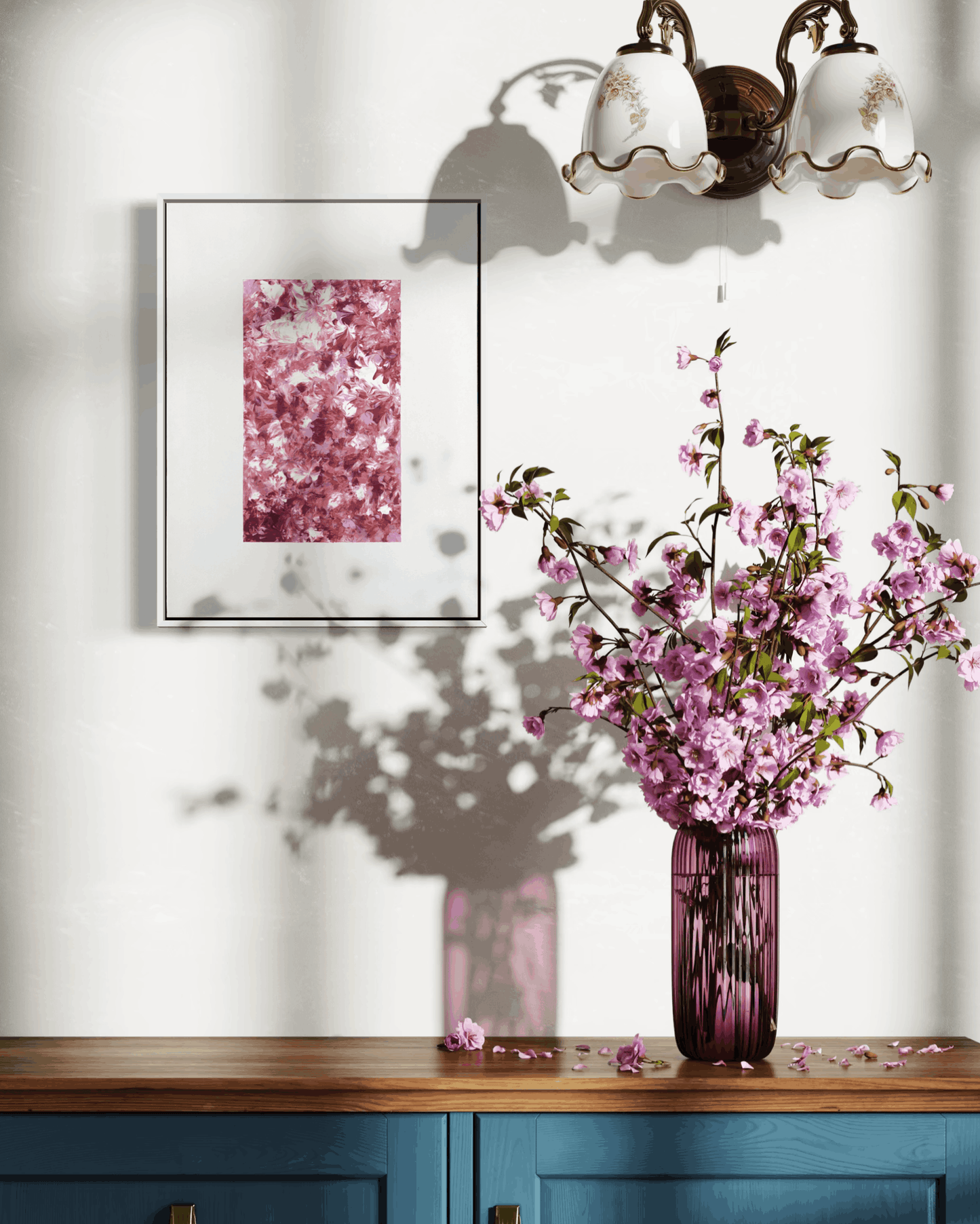 A framed art print featuring a pink cherry blossom pattern, displayed on a white wall above a wooden console table with a blue cabinet base. A matching pink glass vase filled with pink cherry blossom branches sits on the table, casting a shadow on the wall.