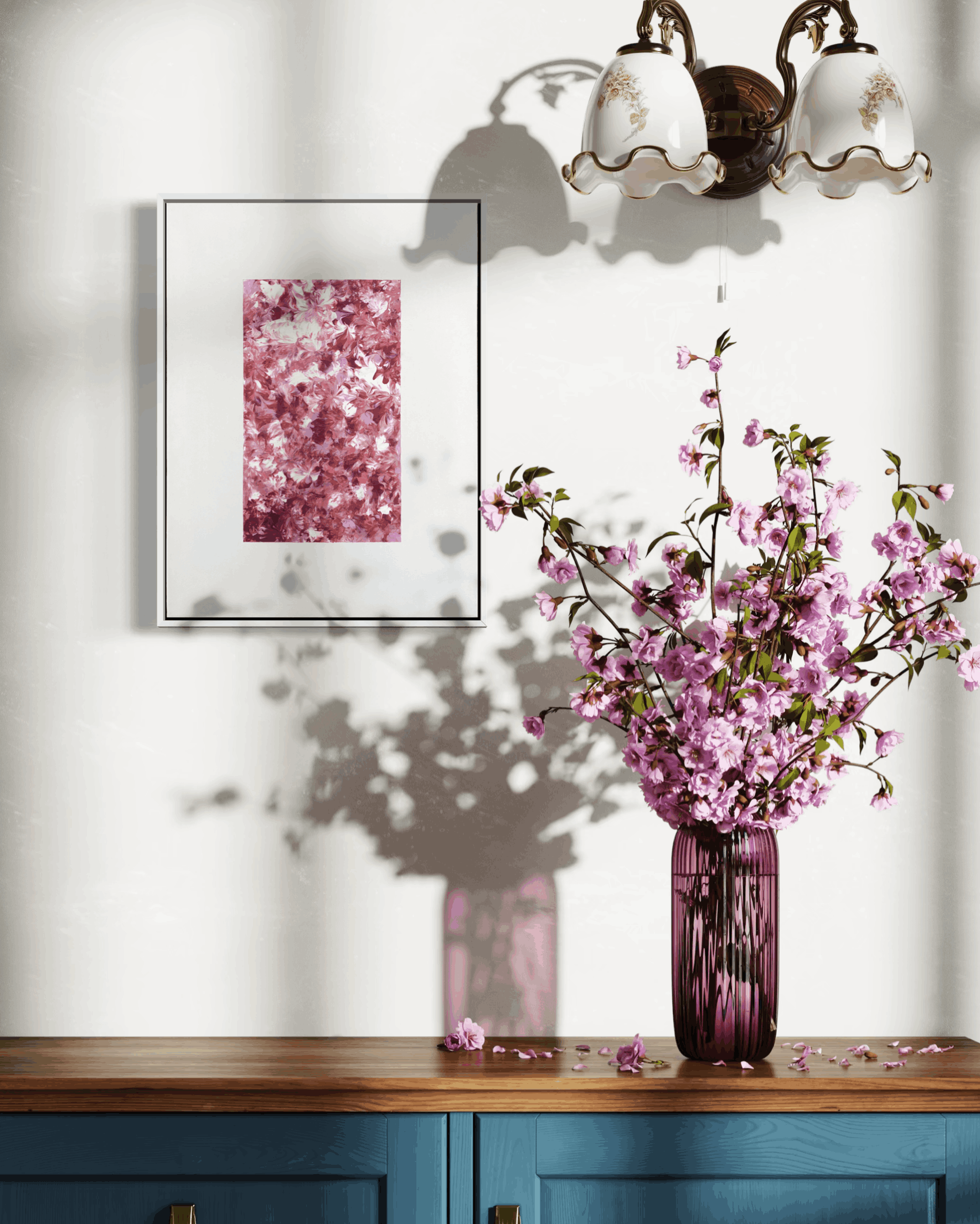 A framed art print featuring a pink cherry blossom pattern, displayed on a white wall above a wooden console table with a blue cabinet base. A matching pink glass vase filled with pink cherry blossom branches sits on the table, casting a shadow on the wall.