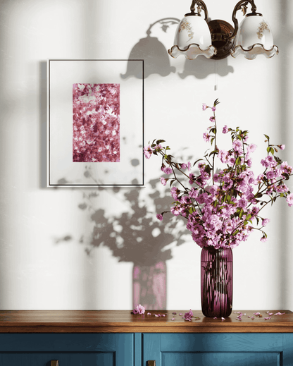 A framed art print featuring a pink cherry blossom pattern, displayed on a white wall above a wooden console table with a blue cabinet base. A matching pink glass vase filled with pink cherry blossom branches sits on the table, casting a shadow on the wall.