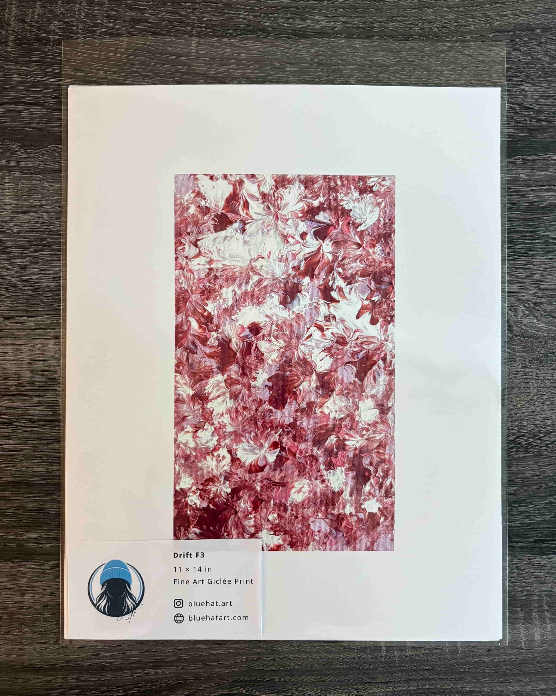 A square art print or card featuring a marbled abstract pattern in pink, red, and white, displayed on a dark wood surface. A small label at the bottom left includes a circular logo and text reading 'Soft Fl' and 'Fine Art Print'.