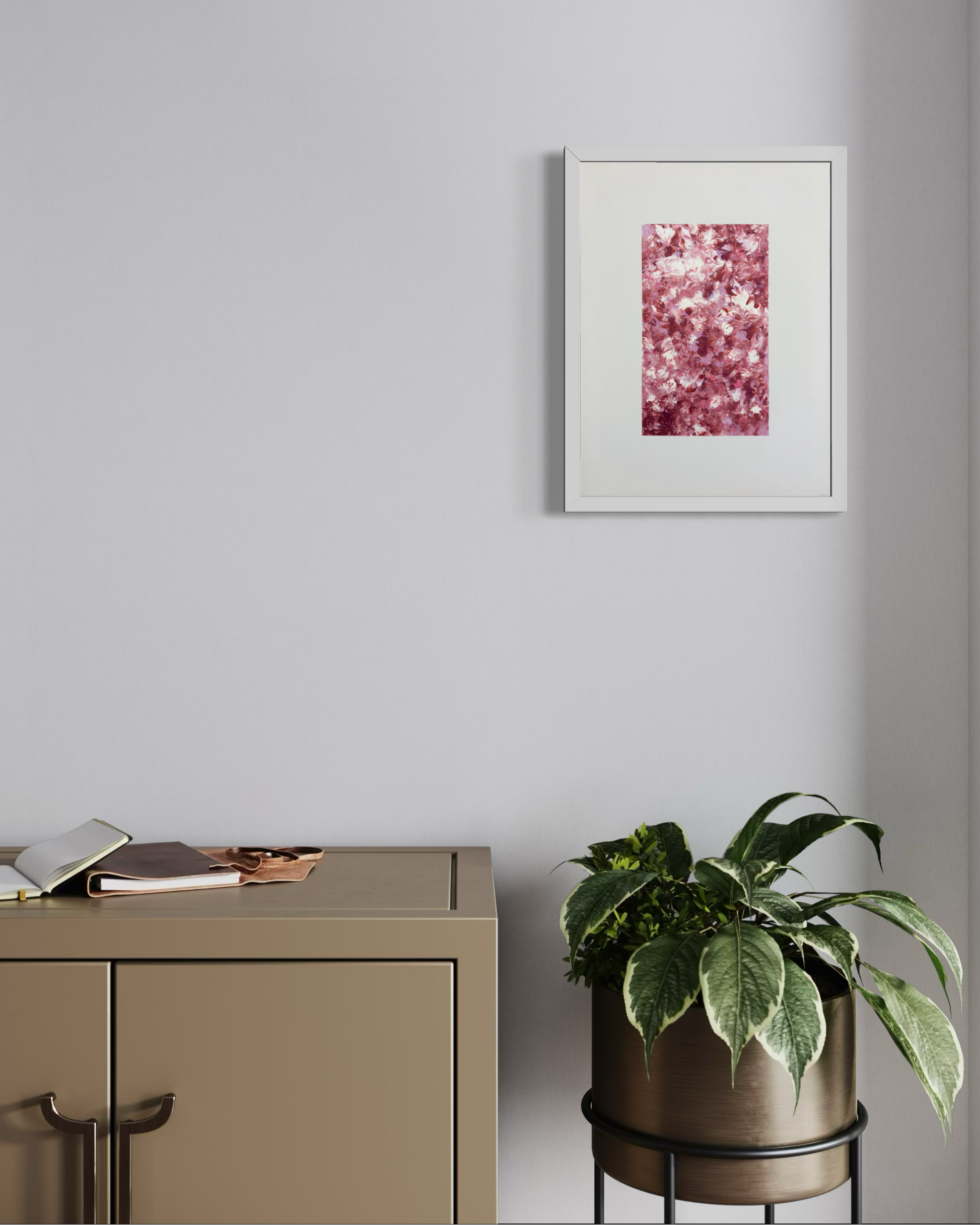 A framed abstract art print with a white frame hangs on a light gray wall above a beige cabinet, next to a potted green plant on a black metal stand.