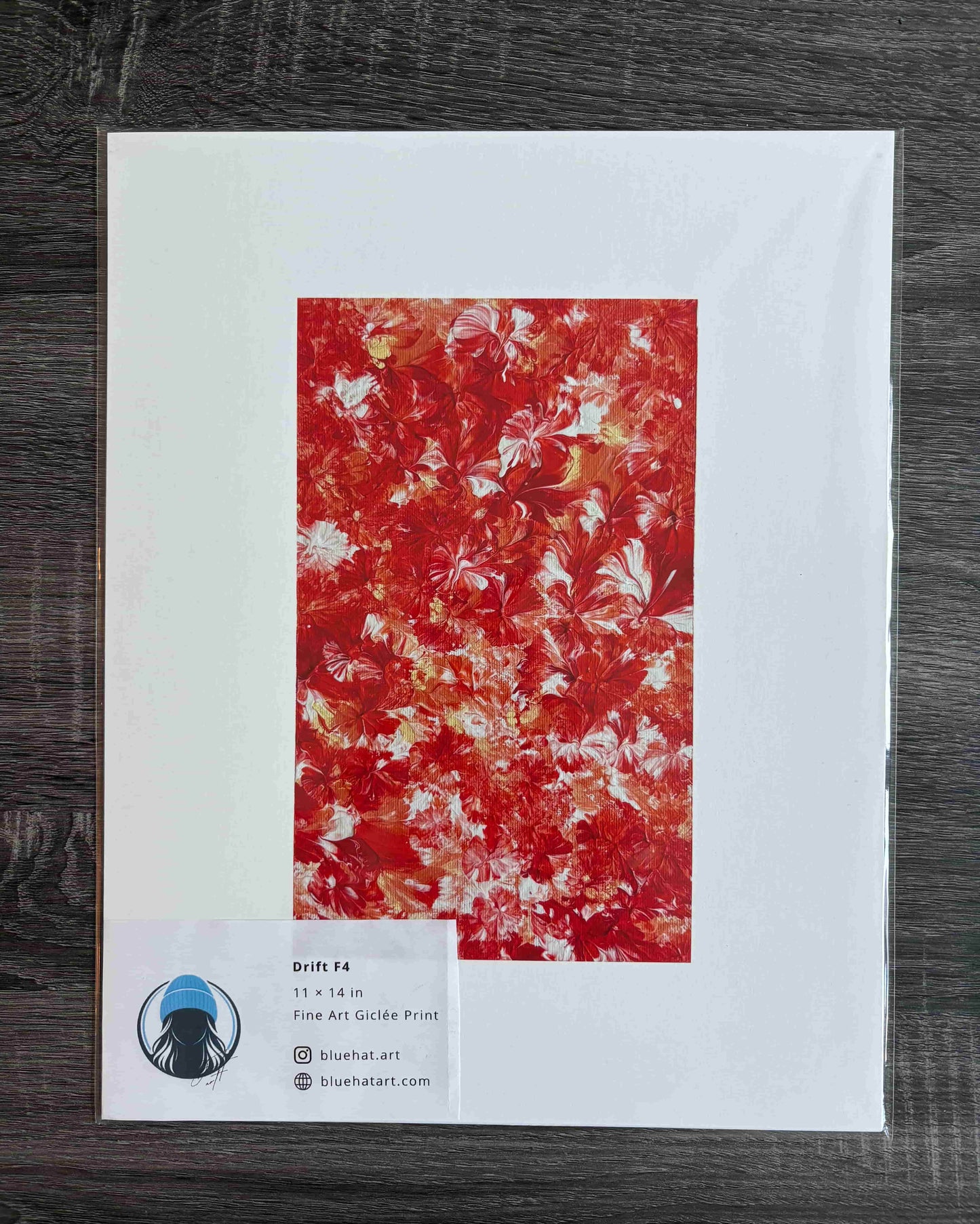 A framed or mounted art print on a white backing card, photographed from above on a dark wood surface. The central artwork is an abstract floral or marbled pattern in vivid red, orange, and cream tones.