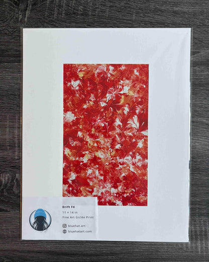 A framed or mounted art print on a white backing card, photographed from above on a dark wood surface. The central artwork is an abstract floral or marbled pattern in vivid red, orange, and cream tones.