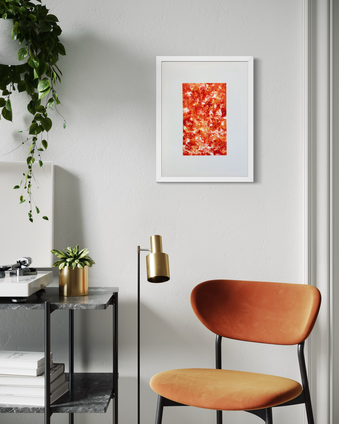 A framed abstract art print featuring a vibrant red and orange textured design, displayed on a light gray wall in a modern room setting with an orange chair, a gold floor lamp, and a potted plant.