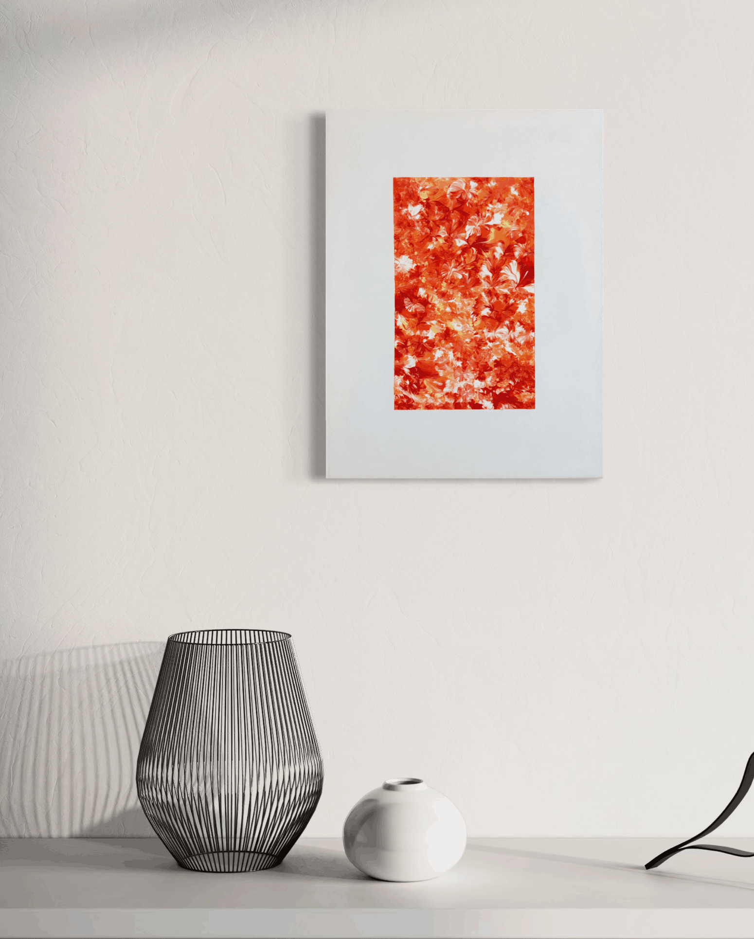A framed abstract art print featuring a vibrant red, orange, and white textured pattern, displayed on a white wall above a minimalist tabletop with a ribbed glass vase and a small white ceramic object.