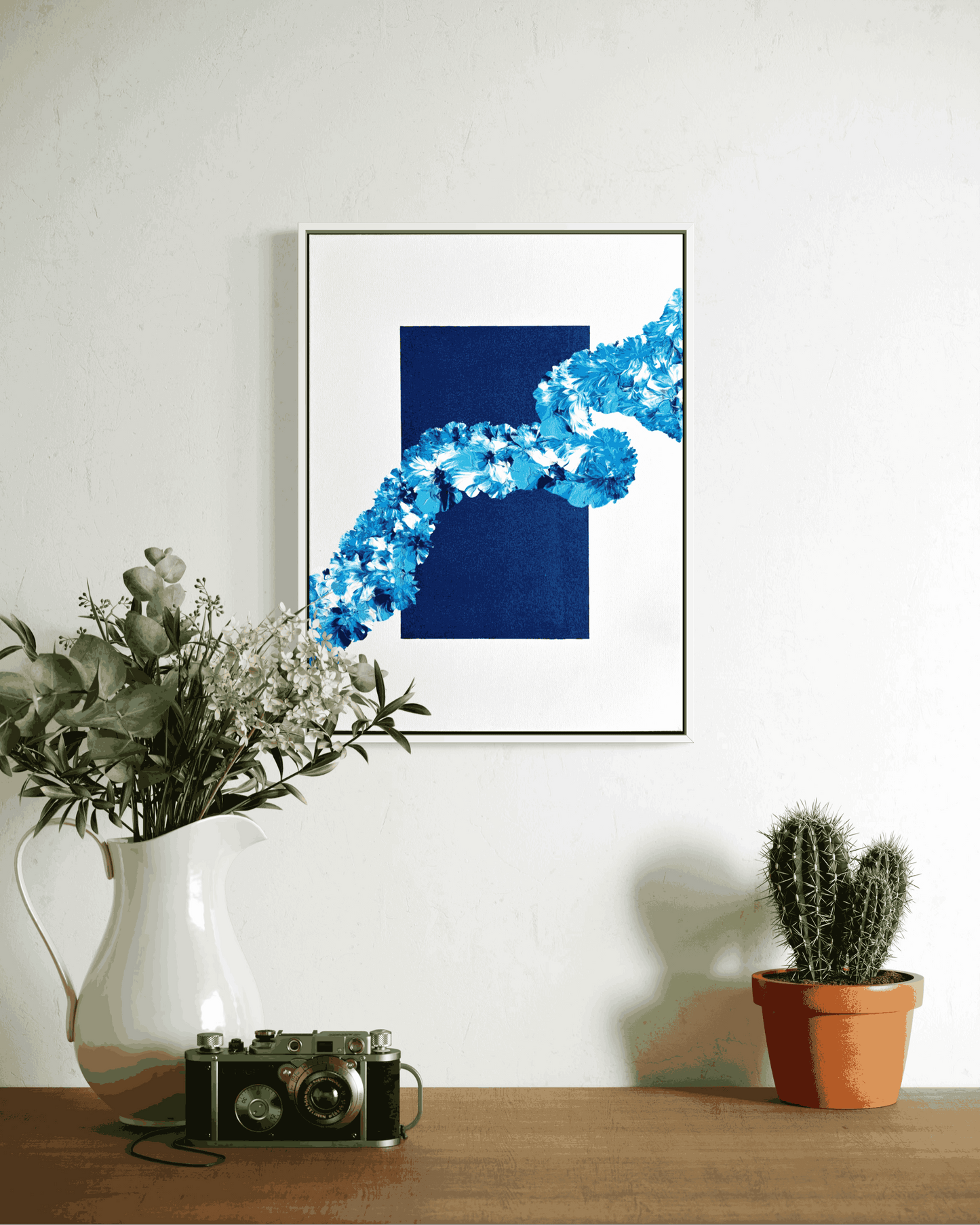 A framed abstract art print featuring a dark blue square and a bright blue floral-like curved pattern, hanging on a white wall above a wooden table with a white vase of eucalyptus, a vintage camera, and a potted cactus.