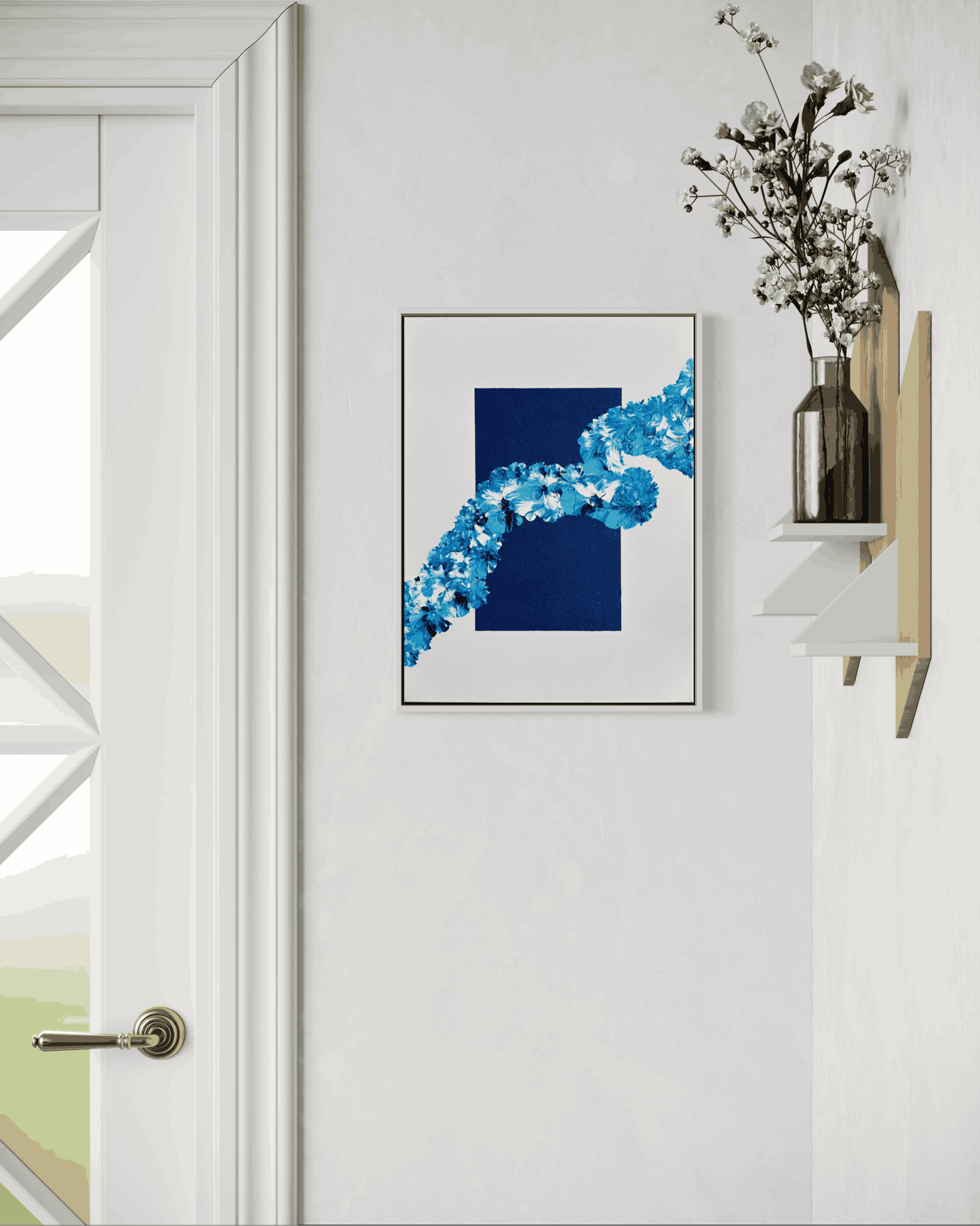 A framed abstract art print featuring a dark navy blue rectangle and a bright blue floral wave pattern, displayed on a white wall next to a wooden shelf with a vase of dried flowers.