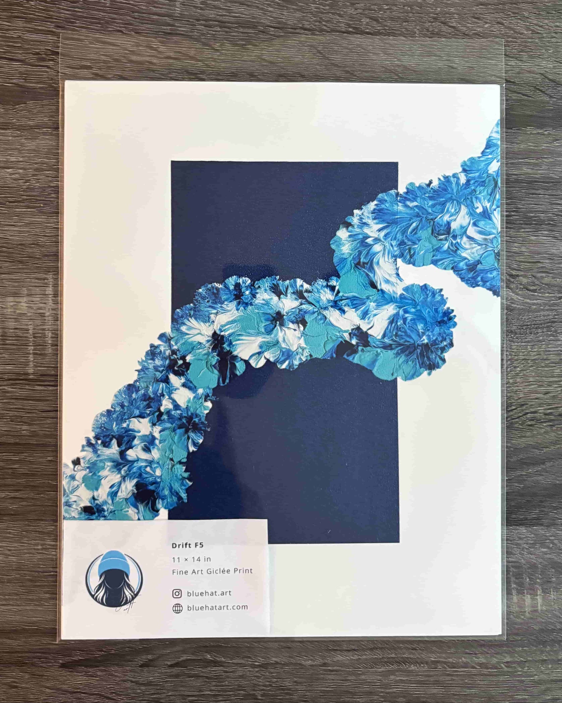 A square art print or poster displayed flat on a dark wood surface, wrapped in a clear protective sleeve. The design features a large dark navy blue square in the center with a flowing, textured blue and white abstract ribbon-like shape entering from the upper right.