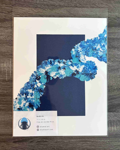 A square art print or poster displayed flat on a dark wood surface, wrapped in a clear protective sleeve. The design features a large dark navy blue square in the center with a flowing, textured blue and white abstract ribbon-like shape entering from the upper right.