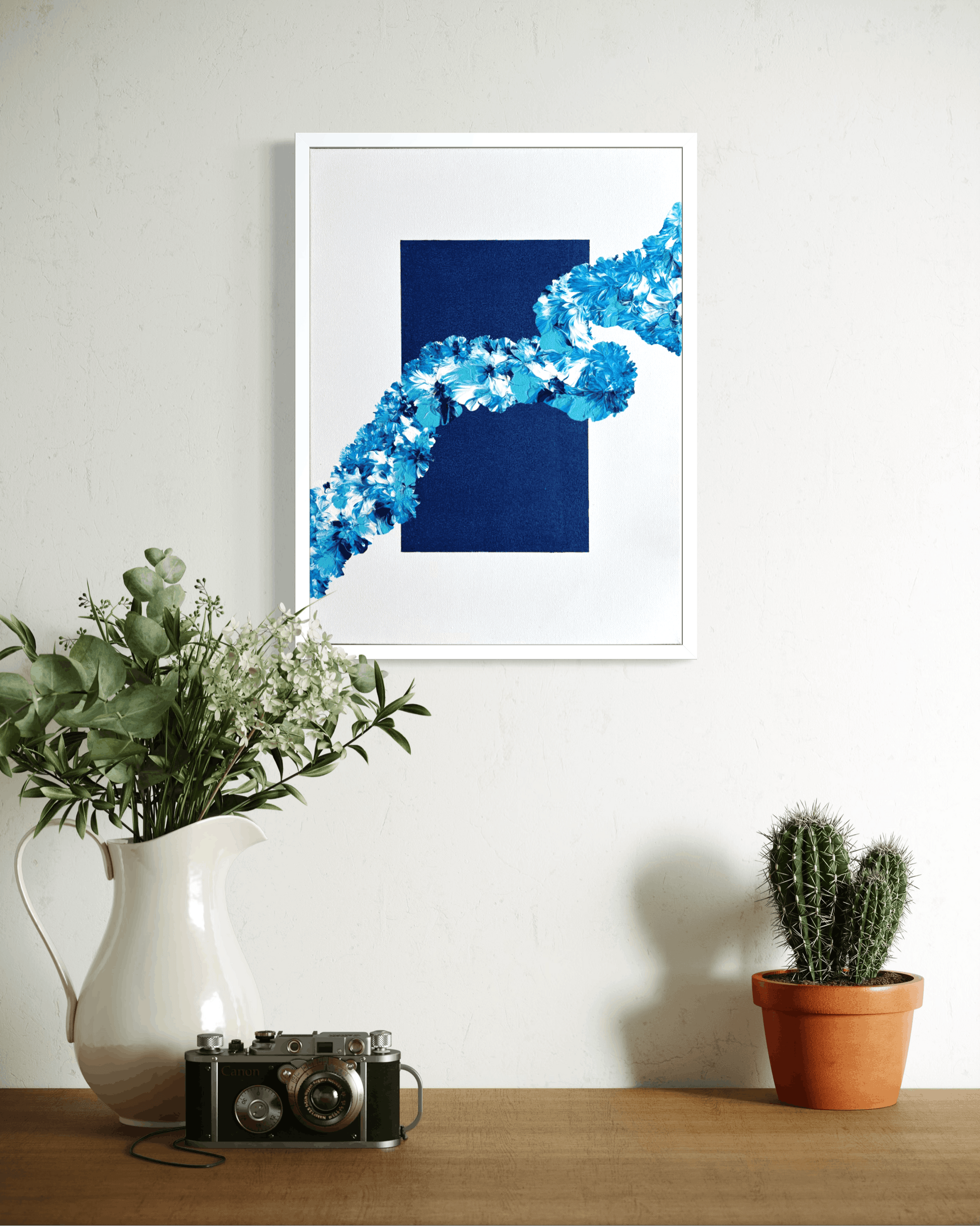 A framed abstract art print featuring a deep navy blue square and a bright blue floral fractal pattern, hanging on a white wall above a wooden table styled with a white pitcher of greenery, a vintage camera, and a potted cactus.