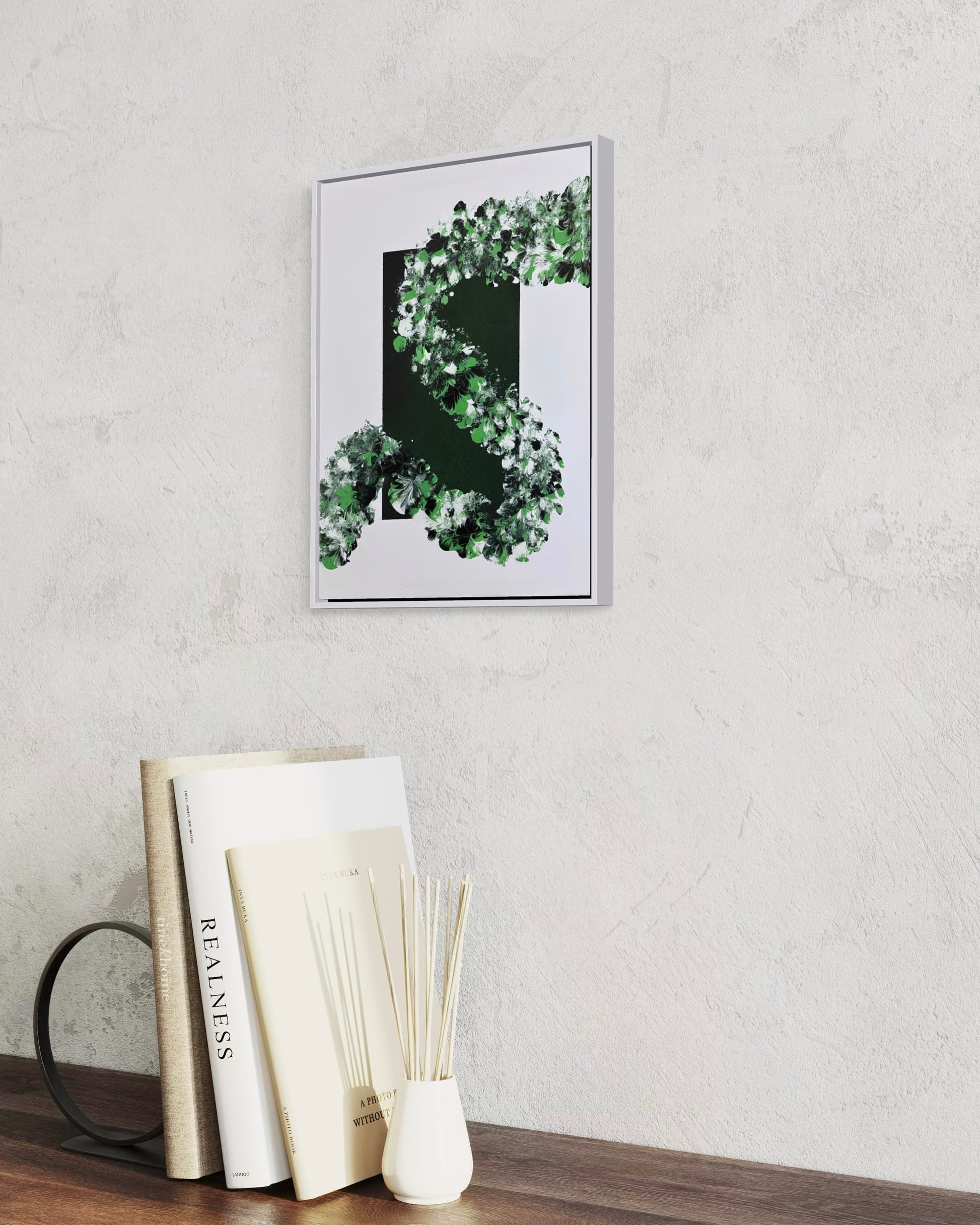 A framed wall art print featuring a green leafy wreath around a dark central rectangle, hanging on a light textured wall above a wooden surface with books and a reed diffuser.