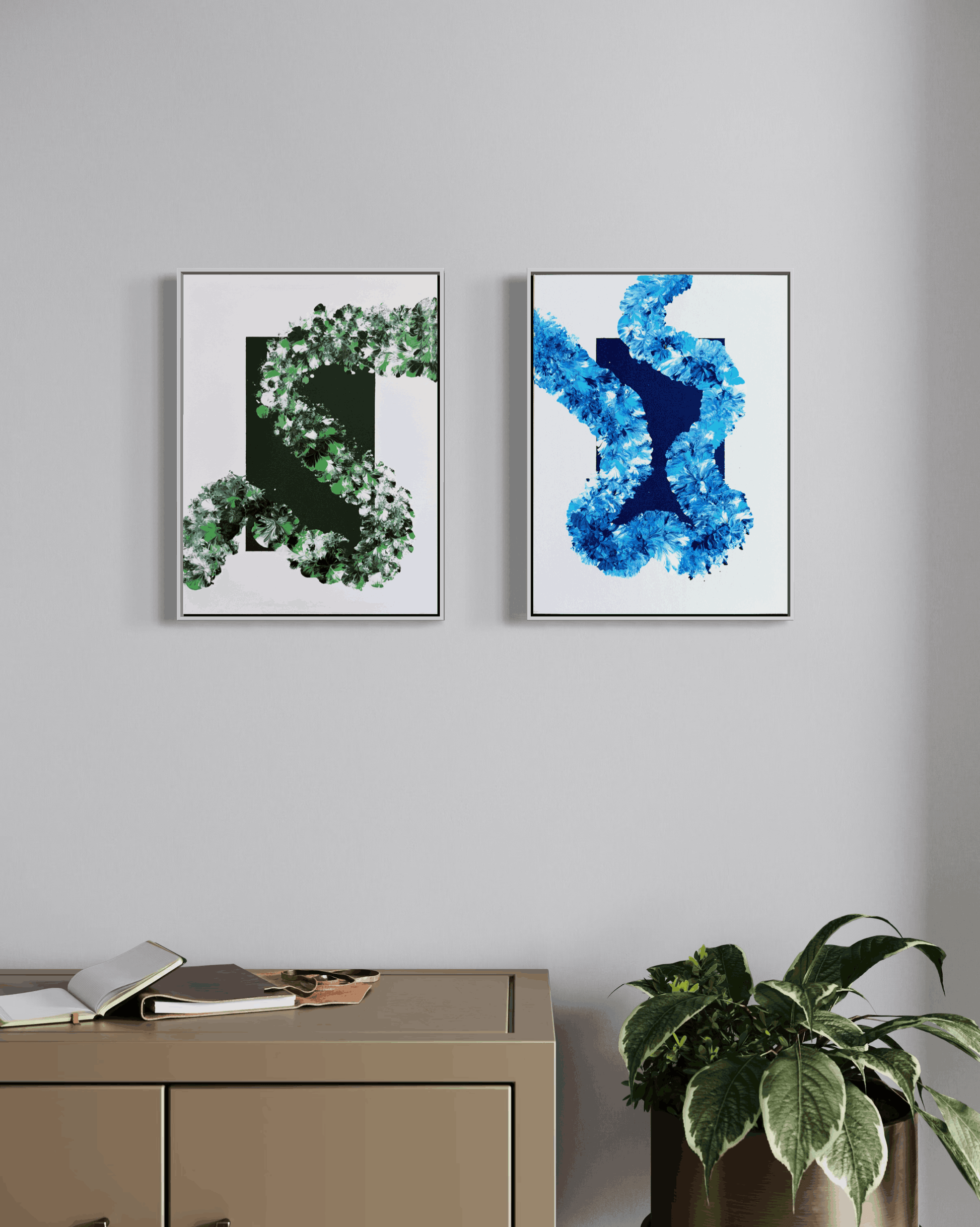 Two framed abstract art prints hanging side-by-side on a light gray wall above a beige cabinet, with a potted plant in the foreground.