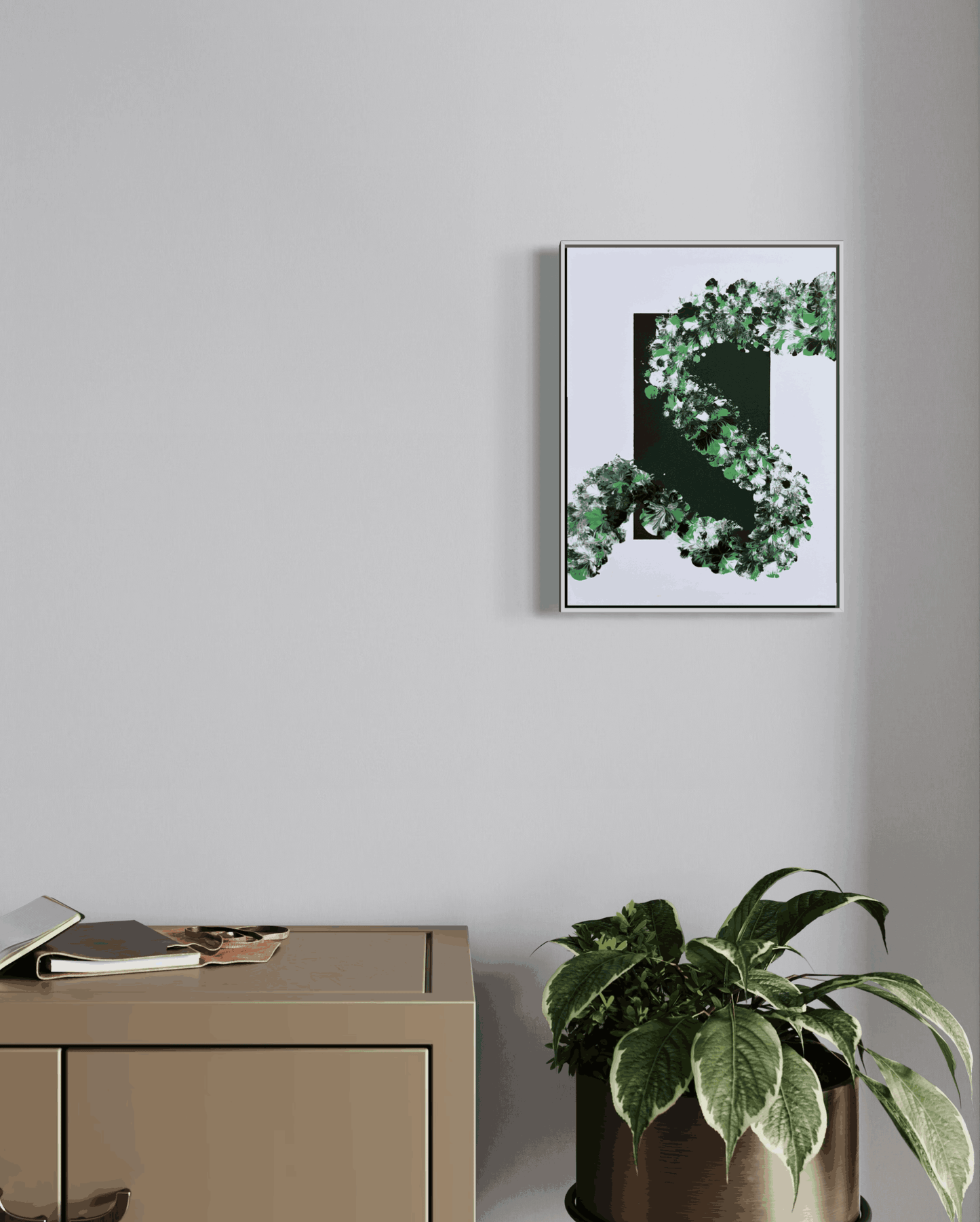 A framed abstract botanical art print featuring green leaves and dark green shapes on a white background, hanging on a light gray wall above a beige cabinet with a potted plant nearby.