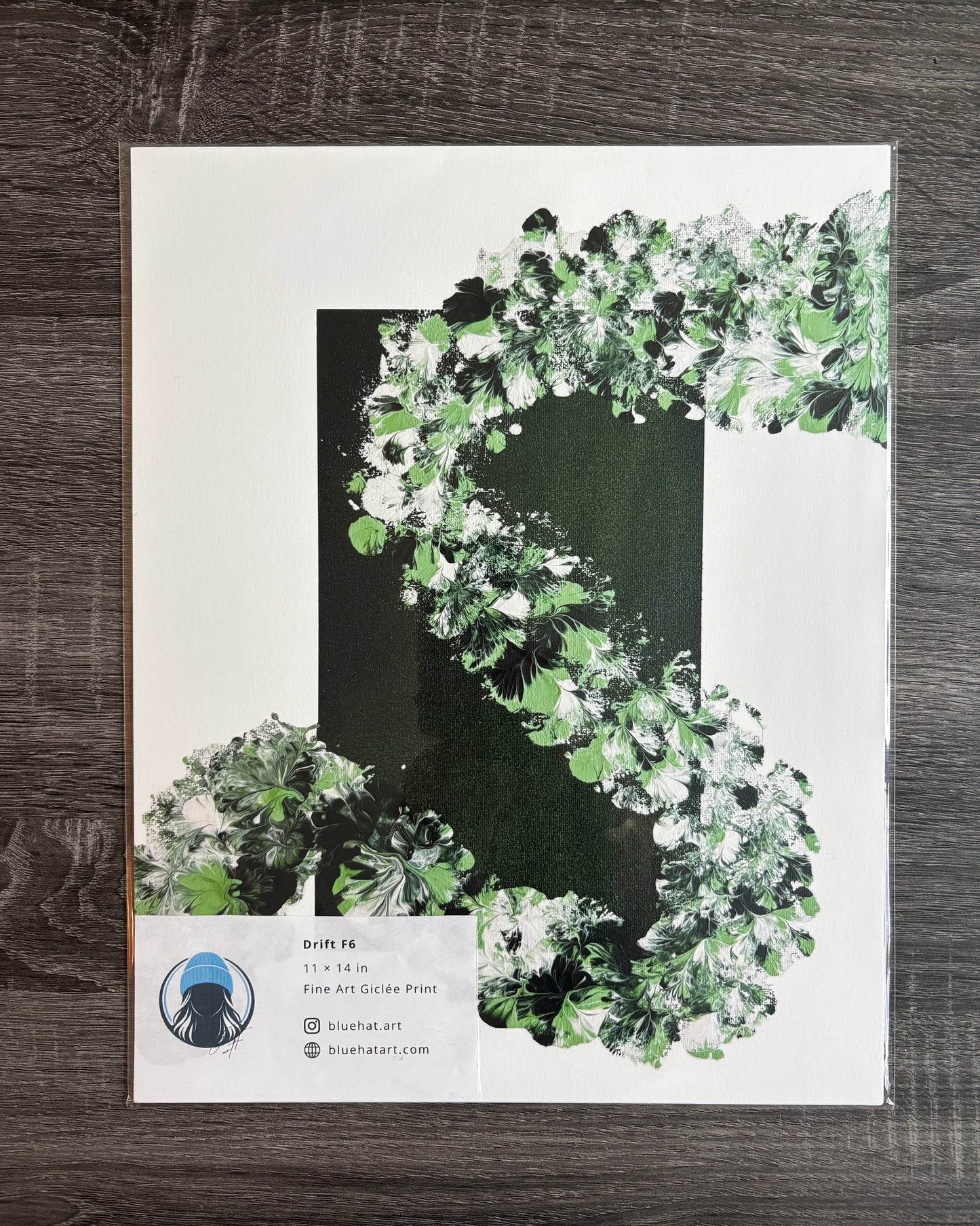 A fine art print featuring a stylized green and white leafy floral design on a white background, displayed flat on a dark wood surface.