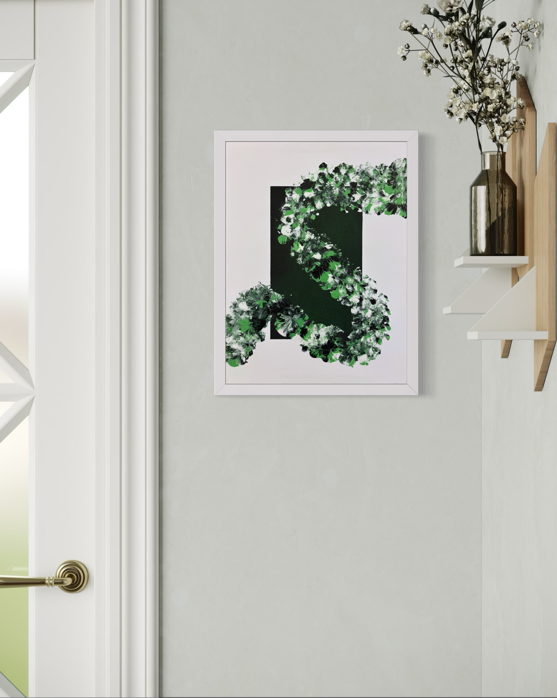 A framed abstract art print featuring a green and white leafy spiral design on a white background, displayed on a light gray wall next to a white door frame and a wooden shelf with a vase of flowers.