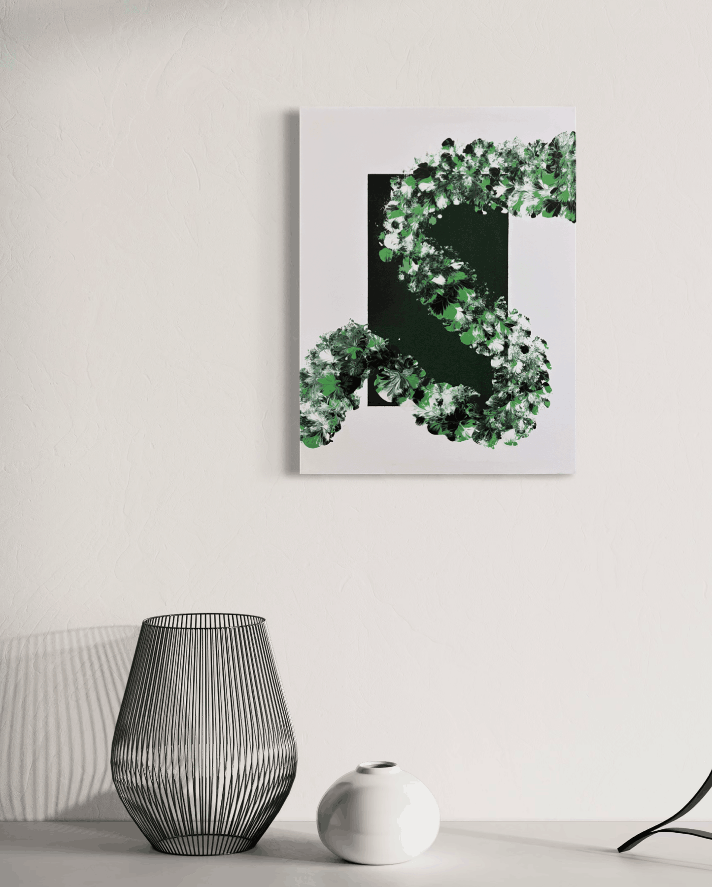 A vertical canvas wall art print featuring a stylized green leaf-like abstract design on a white background, hanging on a light gray wall above a table with a wire lamp and a small white vase.