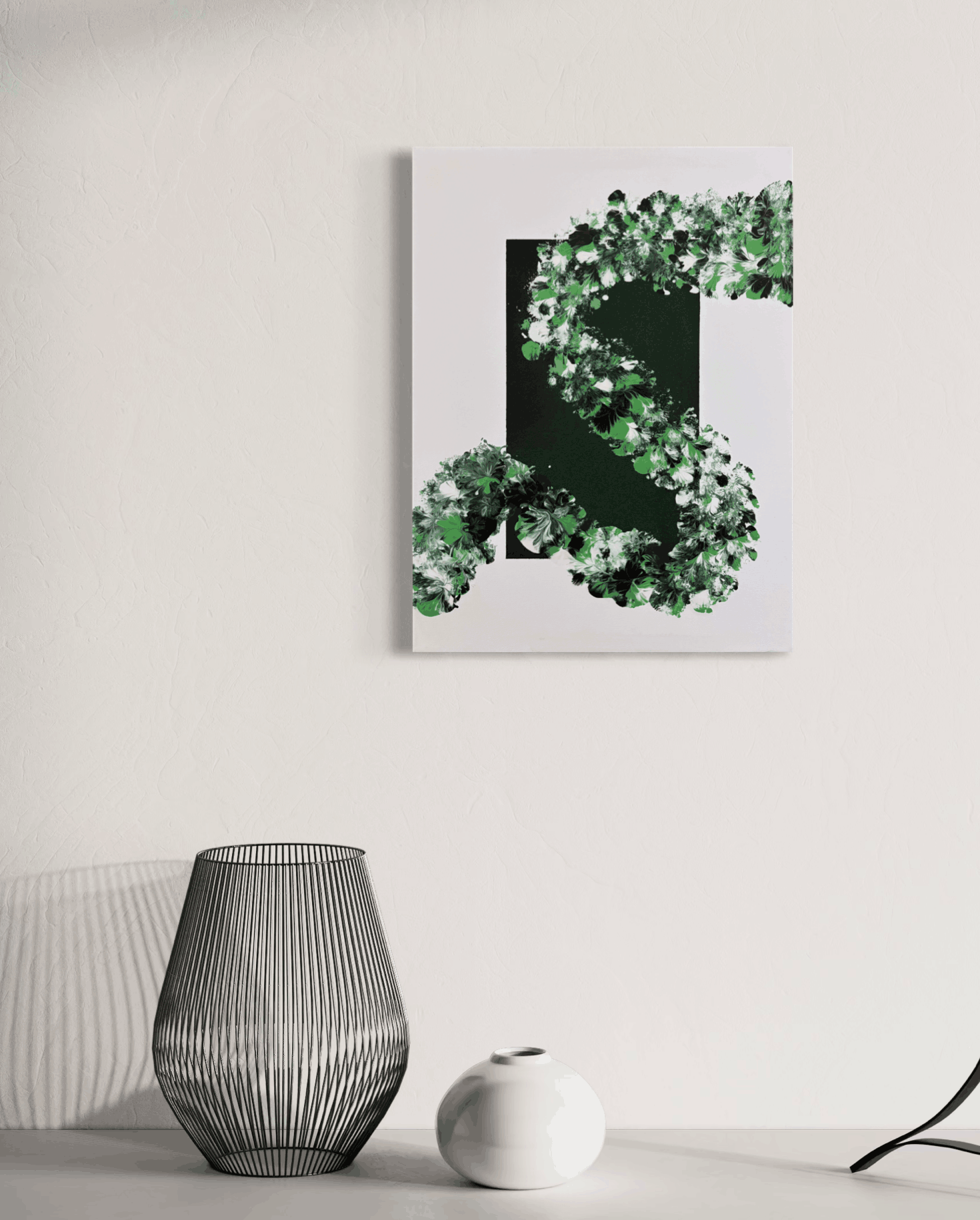 A vertical canvas wall art print featuring a stylized green leaf-like abstract design on a white background, hanging on a light gray wall above a table with a wire lamp and a small white vase.