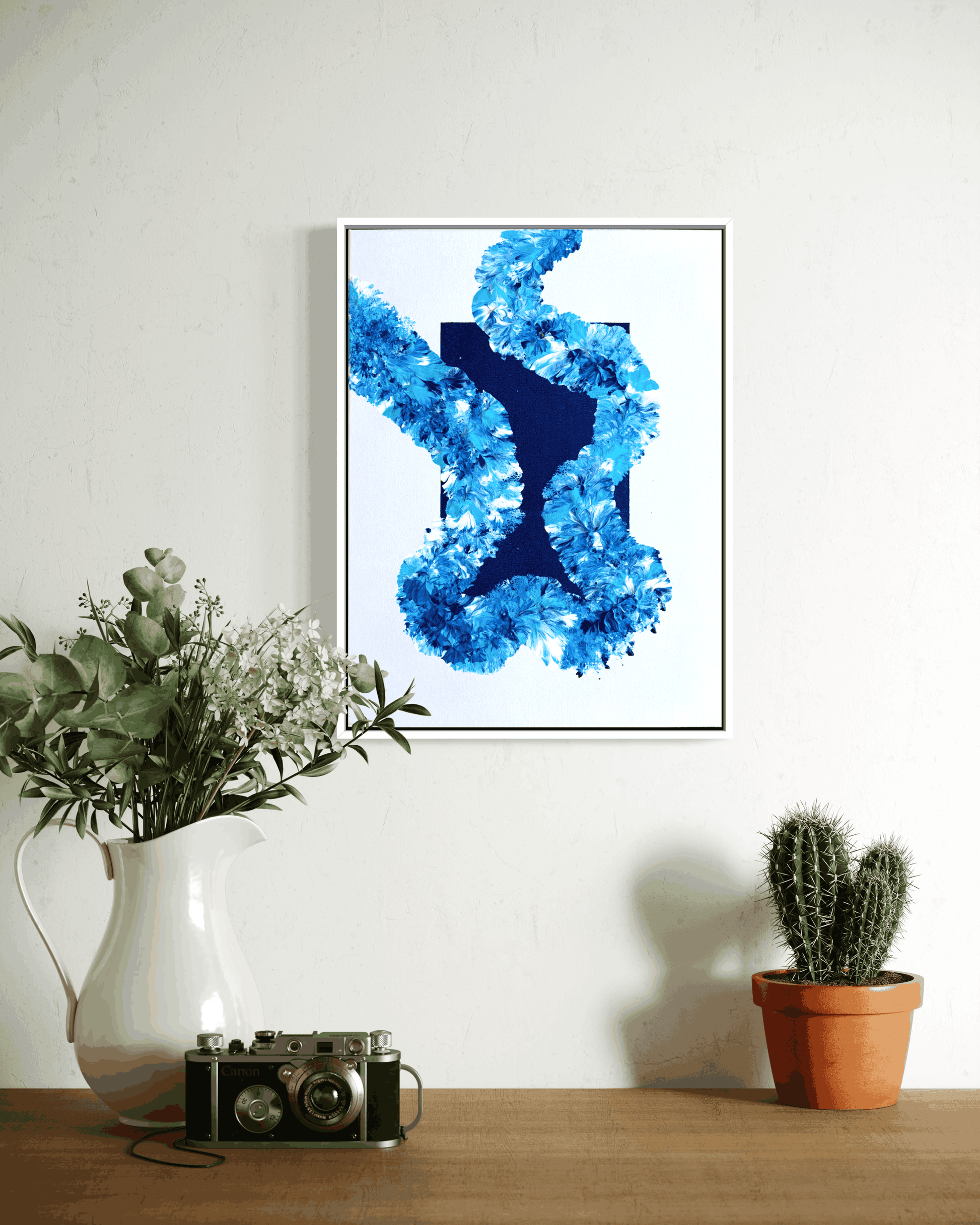 A framed abstract art print featuring a textured blue and navy blue design on a white background, hanging on a white wall above a wooden table styled with a white pitcher of greenery, a vintage camera, and a small potted cactus.